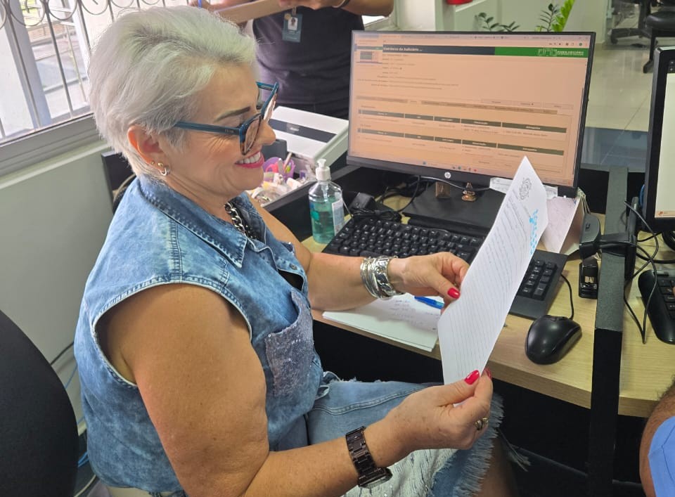 imagem colorida de uma mulher de meia-idade, com cabelos brancos curtos e óculos de aro azul, está sentada em uma mesa de escritório, segurando uma cartinha com as duas mãos e sorrindo.    imagem colorida de uma mulher de meia-idade, com cabelos brancos curtos e óculos de aro azul, está sentada em uma mesa de escritório, segurando uma cartinha com as duas mãos e sorrindo.