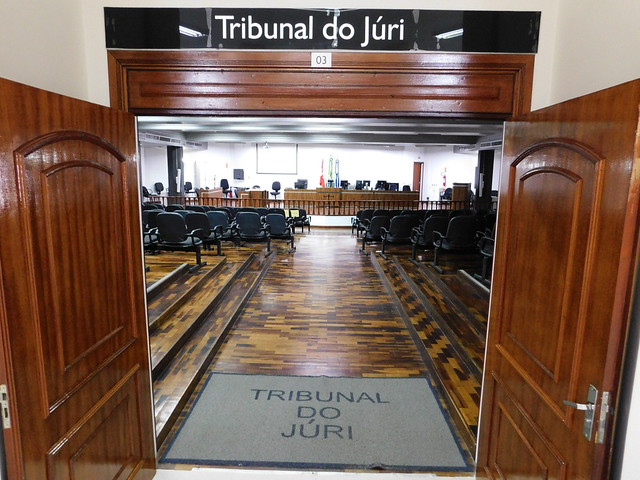 Imagem colorida mostra a entrada de uma sala de audiências de um Tribunal do Júri, com as portas de madeira abertas e um capacho no chão. O interior é visível, com fileiras de cadeiras pretas vazias e a bancada elevada ao fundo.