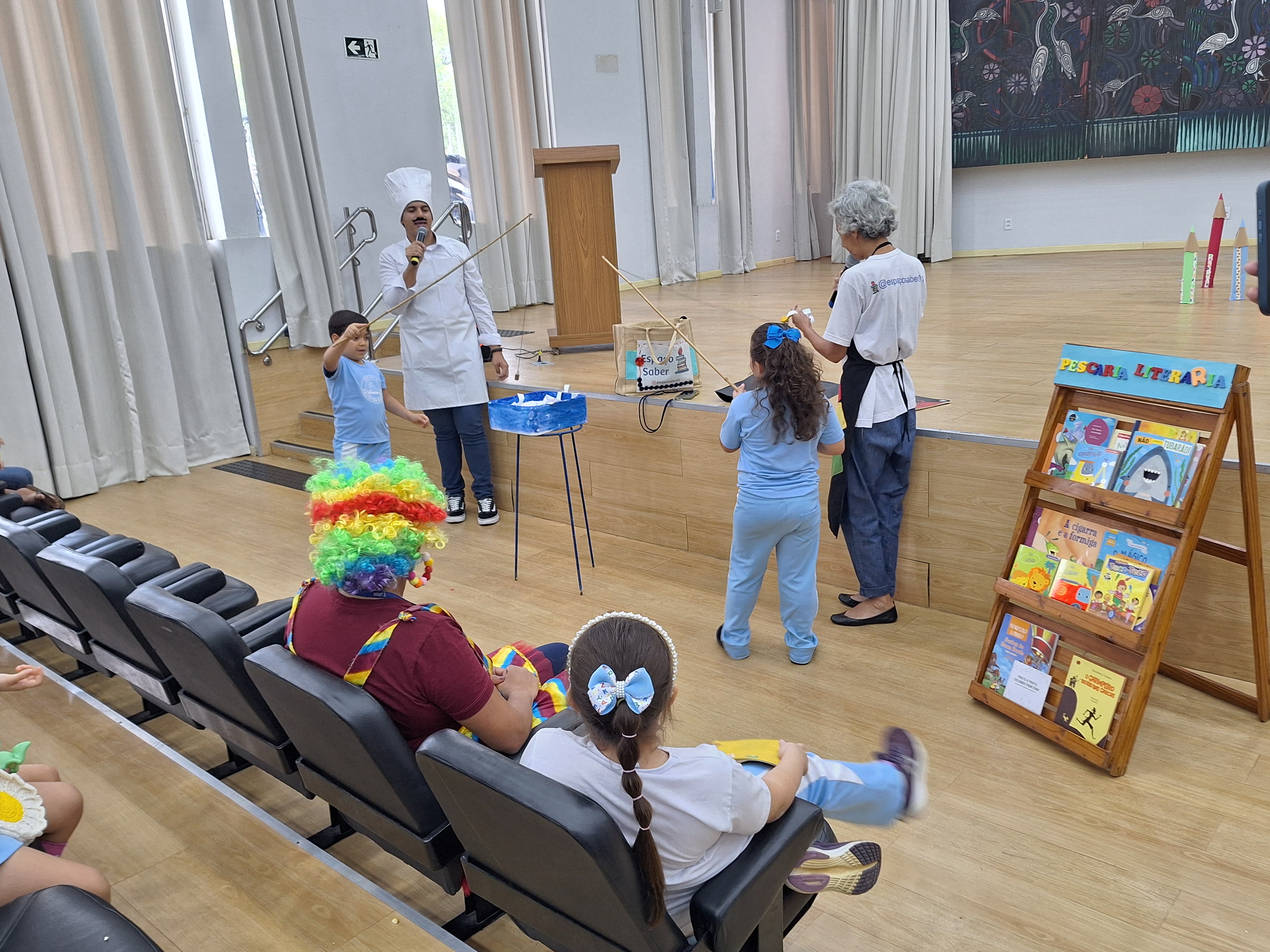 A imagem colorida mostra um auditório com cortinas brancas e piso de madeira, onde ocorre uma atividade infantil chamada “Pesca Literária”. No centro, um homem vestido de chef de cozinha, com avental e chapéu branco, segura um microfone e orienta duas crianças que participam da brincadeira de pesca usando varas com anzóis de brinquedo. Ao lado deles, uma mulher de cabelos grisalhos, também de avental, ajuda na atividade. Em primeiro plano, há uma pessoa vestida de palhaço, com peruca colorida, sentada nas cadeiras da plateia, observando. Perto do palco, um expositor de madeira exibe livros infantis coloridos. Crianças com uniformes azuis acompanham a ação com atenção e alegria. A imagem colorida mostra um auditório com cortinas brancas e piso de madeira, onde ocorre uma atividade infantil chamada “Pesca Literária”. No centro, um homem vestido de chef de cozinha, com avental e chapéu branco, segura um microfone e orienta duas crianças que participam da brincadeira de pesca usando varas com anzóis de brinquedo. Ao lado deles, uma mulher de cabelos grisalhos, também de avental, ajuda na atividade. Em primeiro plano, há uma pessoa vestida de palhaço, com peruca colorida, sentada nas cadeiras da plateia, observando. Perto do palco, um expositor de madeira exibe livros infantis coloridos. Crianças com uniformes azuis acompanham a ação com atenção e alegria.