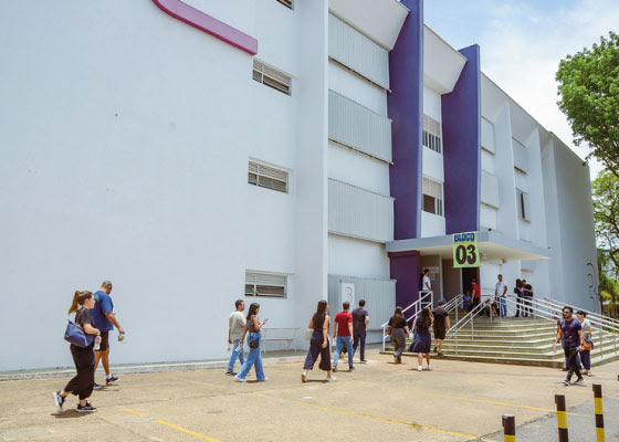 A imagem mostra o exterior de um prédio de universidade branco e azul, com a inscrição BLOCO 03 sobre a entrada. Várias pessoas, possivelmente estudantes, estão andando em direção ou saindo do prédio. A imagem mostra o exterior de um prédio de universidade branco e azul, com a inscrição BLOCO 03 sobre a entrada. Várias pessoas, possivelmente estudantes, estão andando em direção ou saindo do prédio.