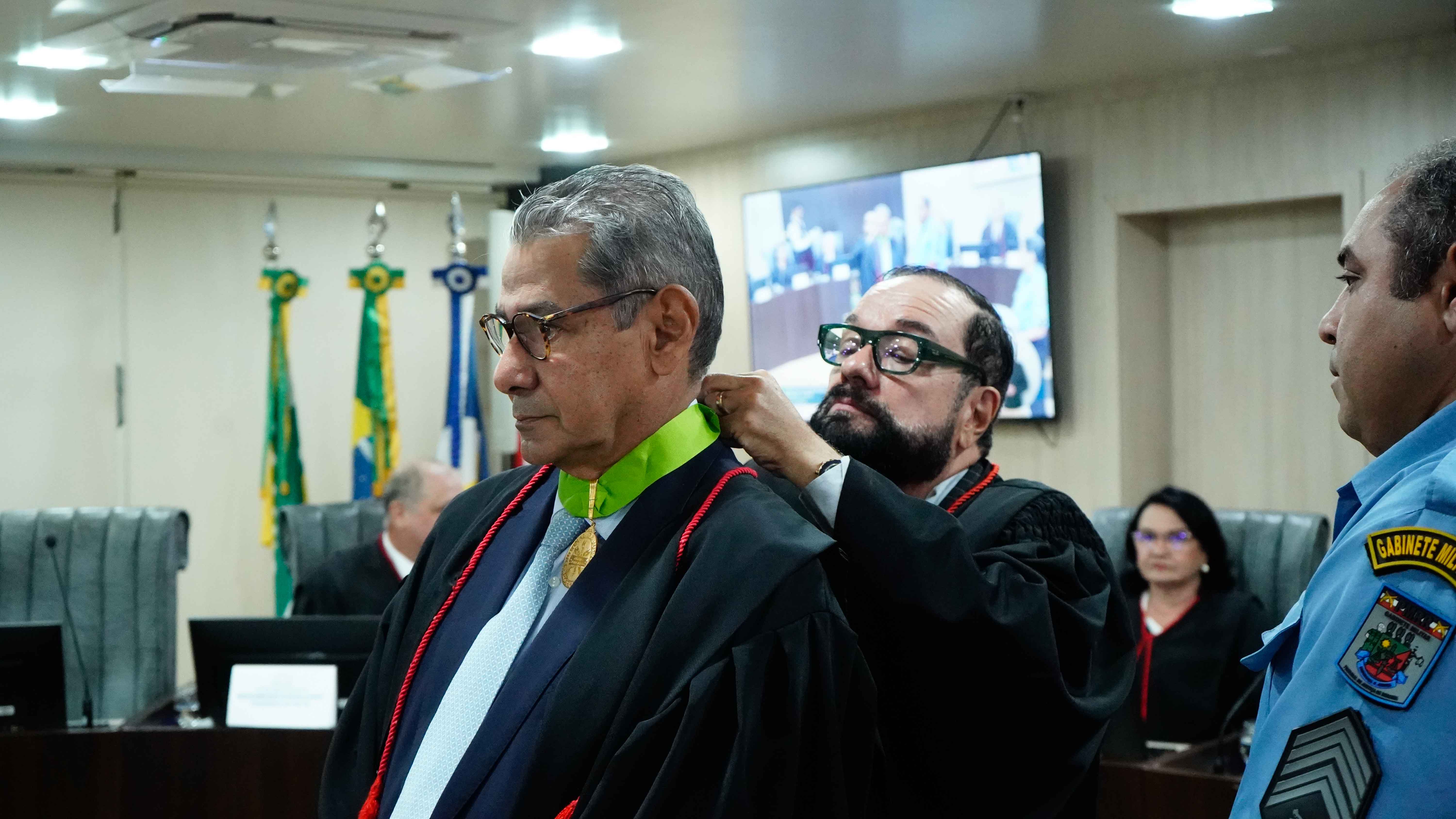Imagem colorida mostra em cerimônia solene, o presidente do TJRR, Desembargador Leonardo Cupello, ajusta um colar de comenda no Procurador de Justiça Sales Eurico Melgarejo Freitas. Ambos estão de toga em um plenário.  Imagem colorida mostra em cerimônia solene, o presidente do TJRR, Desembargador Leonardo Cupello, ajusta um colar de comenda no Procurador de Justiça Sales Eurico Melgarejo Freitas. Ambos estão de toga em um plenário.