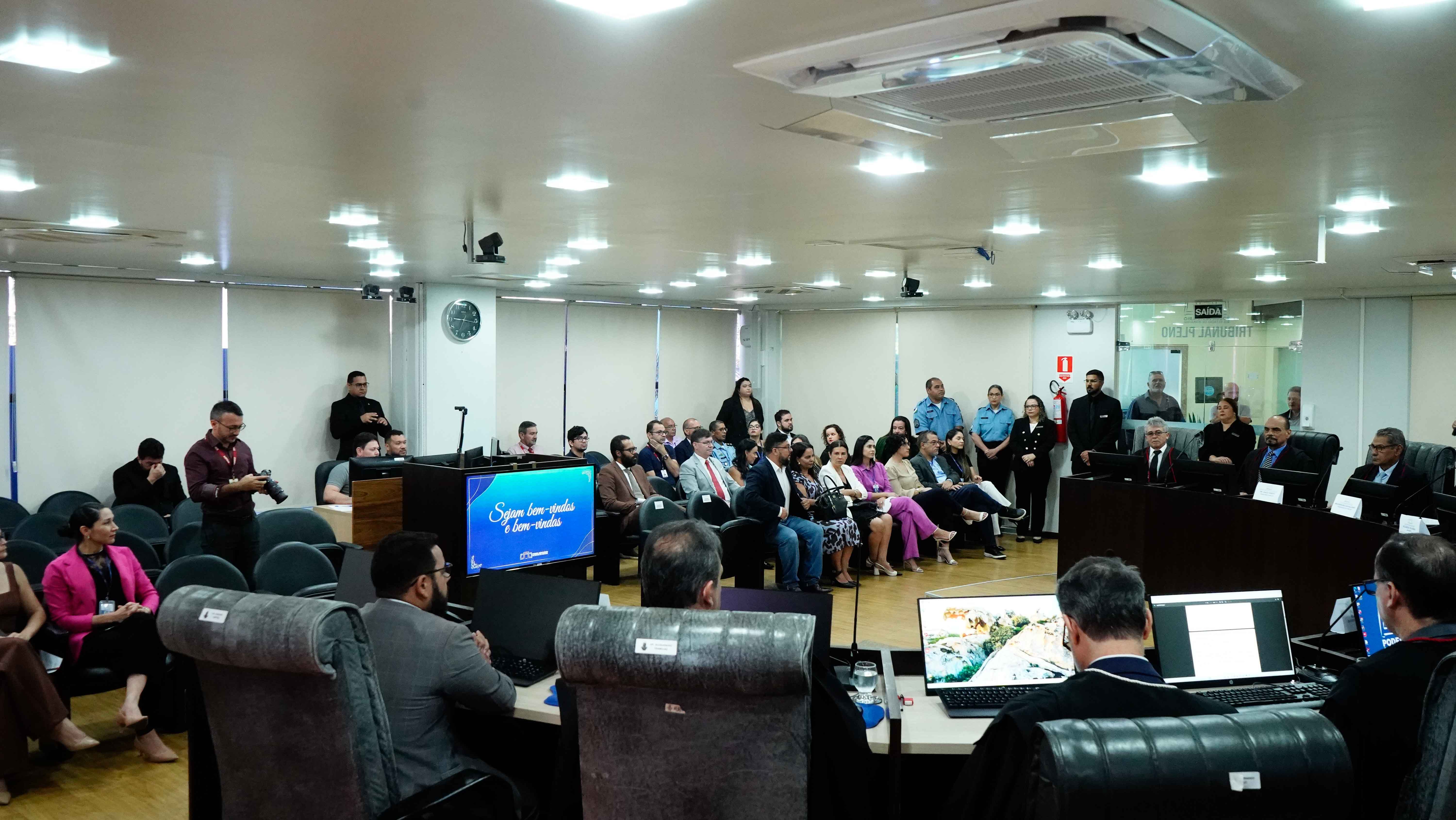 Imagem colorida mostra vista ampla de um plenário lotado durante um evento, com a foto tirada por trás da bancada principal. Em primeiro plano, veem-se as costas de autoridades sentadas em suas cadeiras, e, ao fundo, o público que assiste à cerimônia, sentado e em pé.  Imagem colorida mostra vista ampla de um plenário lotado durante um evento, com a foto tirada por trás da bancada principal. Em primeiro plano, veem-se as costas de autoridades sentadas em suas cadeiras, e, ao fundo, o público que assiste à cerimônia, sentado e em pé.