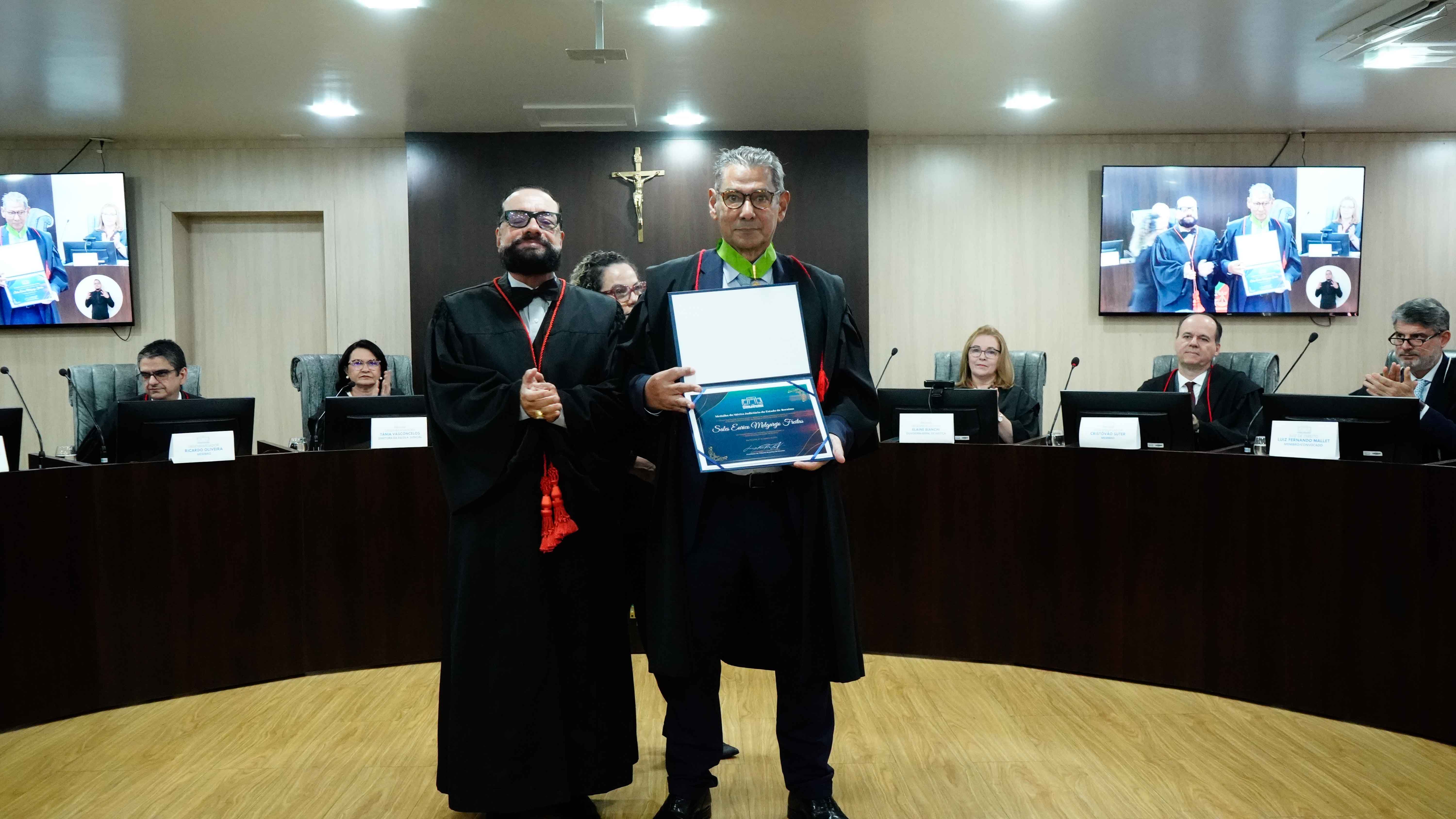Imagem colorida mostra em um plenário, o Procurador de Justiça Sales Eurico Melgarejo Freitas, já condecorado com a comenda, exibe um certificado emoldurado. Ao seu lado, posa o presidente do TJRR, Desembargador Leonardo Cupello. Ambos estão de toga, em frente à bancada das autoridades. Imagem colorida mostra em um plenário, o Procurador de Justiça Sales Eurico Melgarejo Freitas, já condecorado com a comenda, exibe um certificado emoldurado. Ao seu lado, posa o presidente do TJRR, Desembargador Leonardo Cupello. Ambos estão de toga, em frente à bancada das autoridades.