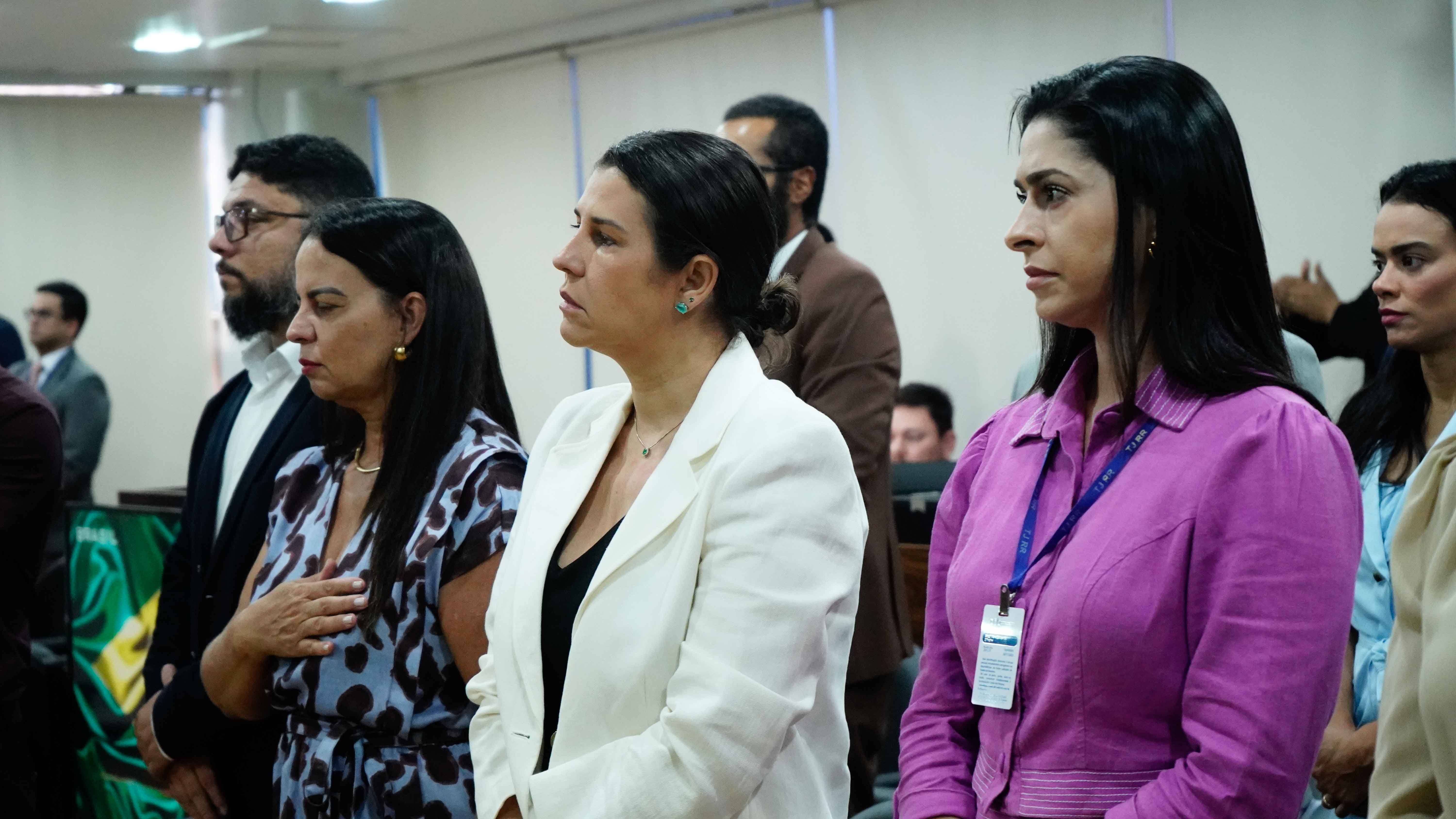 Imagem colorida mostra um grupo de pessoas assiste a uma cerimônia, em pé e de perfil. Em primeiro plano, três mulheres e um homem estão alinhados, olhando atentamente para a esquerda. Uma mulher de vestido estampado está com a mão sobre o peito, ao lado de outra de blazer branco e uma terceira de blazer roxo.  Imagem colorida mostra um grupo de pessoas assiste a uma cerimônia, em pé e de perfil. Em primeiro plano, três mulheres e um homem estão alinhados, olhando atentamente para a esquerda. Uma mulher de vestido estampado está com a mão sobre o peito, ao lado de outra de blazer branco e uma terceira de blazer roxo.