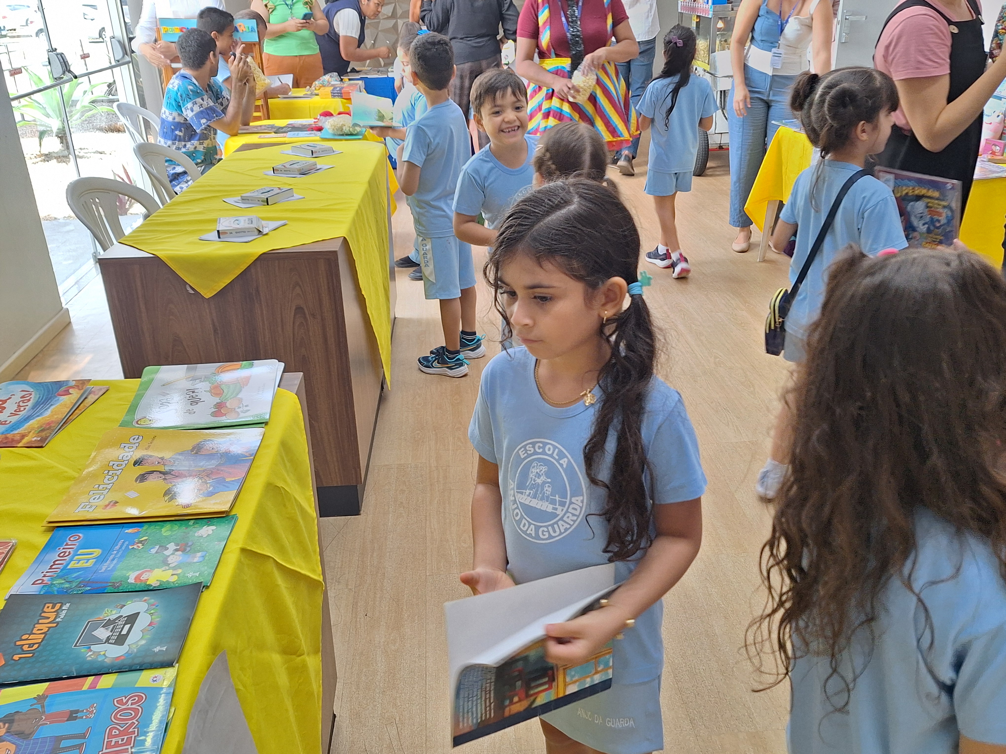 A imagem colorida mostra crianças com uniformes azuis enquanto participam de uma feira de livros em um espaço iluminado por luz natural. Mesas cobertas com toalhas amarelas exibem livros infantis coloridos e materiais educativos. Algumas crianças observam as mesas com curiosidade, enquanto outras folheiam livros. Ao fundo, adultos e monitores interagem com os pequenos e acompanham as atividades. O ambiente é alegre e educativo, com clima de descoberta e aprendizado. A imagem colorida mostra crianças com uniformes azuis enquanto participam de uma feira de livros em um espaço iluminado por luz natural. Mesas cobertas com toalhas amarelas exibem livros infantis coloridos e materiais educativos. Algumas crianças observam as mesas com curiosidade, enquanto outras folheiam livros. Ao fundo, adultos e monitores interagem com os pequenos e acompanham as atividades. O ambiente é alegre e educativo, com clima de descoberta e aprendizado.