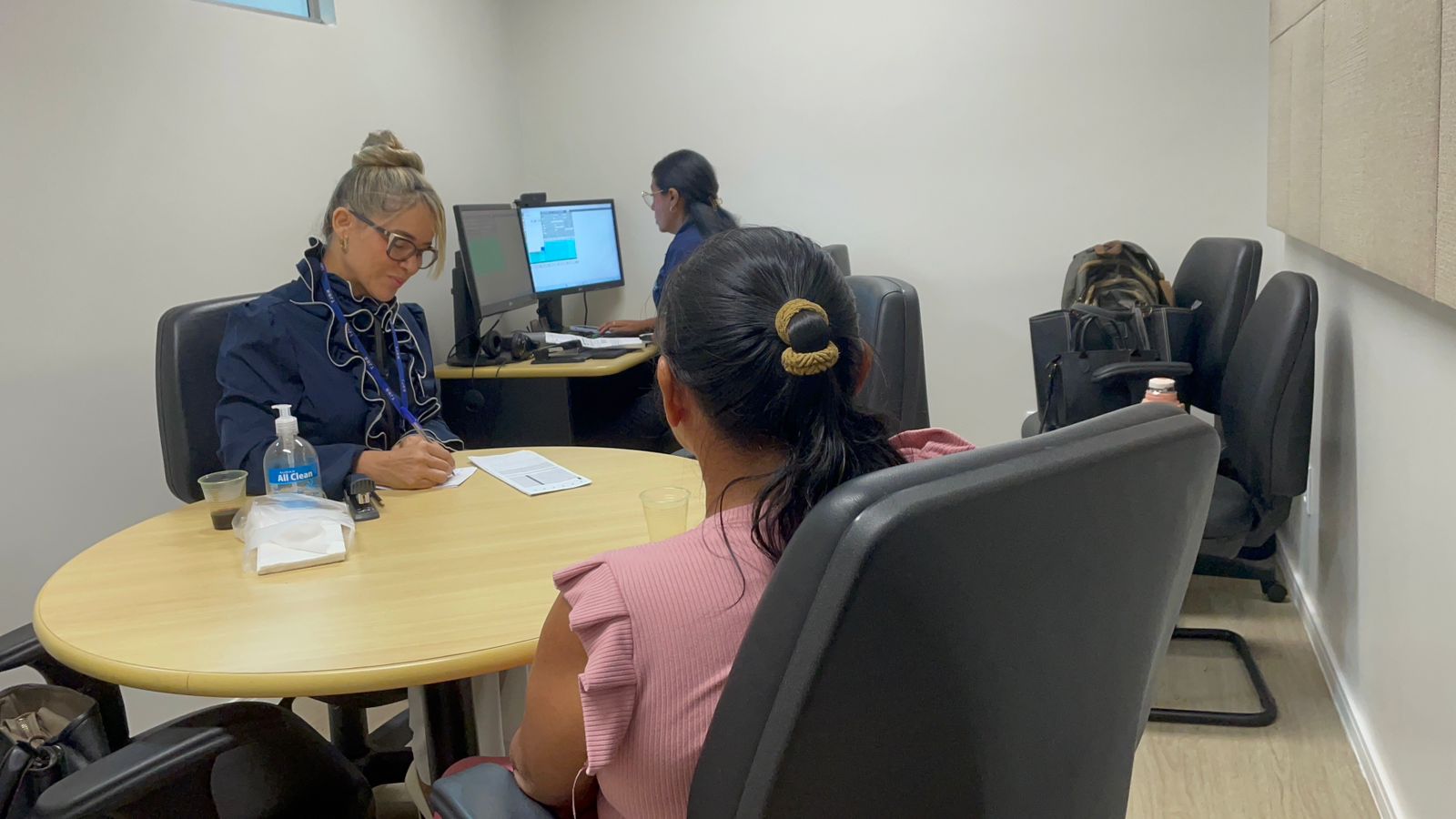 A foto mostra uma sala pequena e clara, com três pessoas reunidas em torno de uma mesa redonda de madeira. Dois homens conversam e fazem anotações, enquanto uma mulher, ao fundo, trabalha em um computador com dois monitores. O ambiente tem cadeiras pretas giratórias, paredes brancas e painéis acústicos bege. A foto mostra uma sala pequena e clara, com três pessoas reunidas em torno de uma mesa redonda de madeira. Dois homens conversam e fazem anotações, enquanto uma mulher, ao fundo, trabalha em um computador com dois monitores. O ambiente tem cadeiras pretas giratórias, paredes brancas e painéis acústicos bege.
