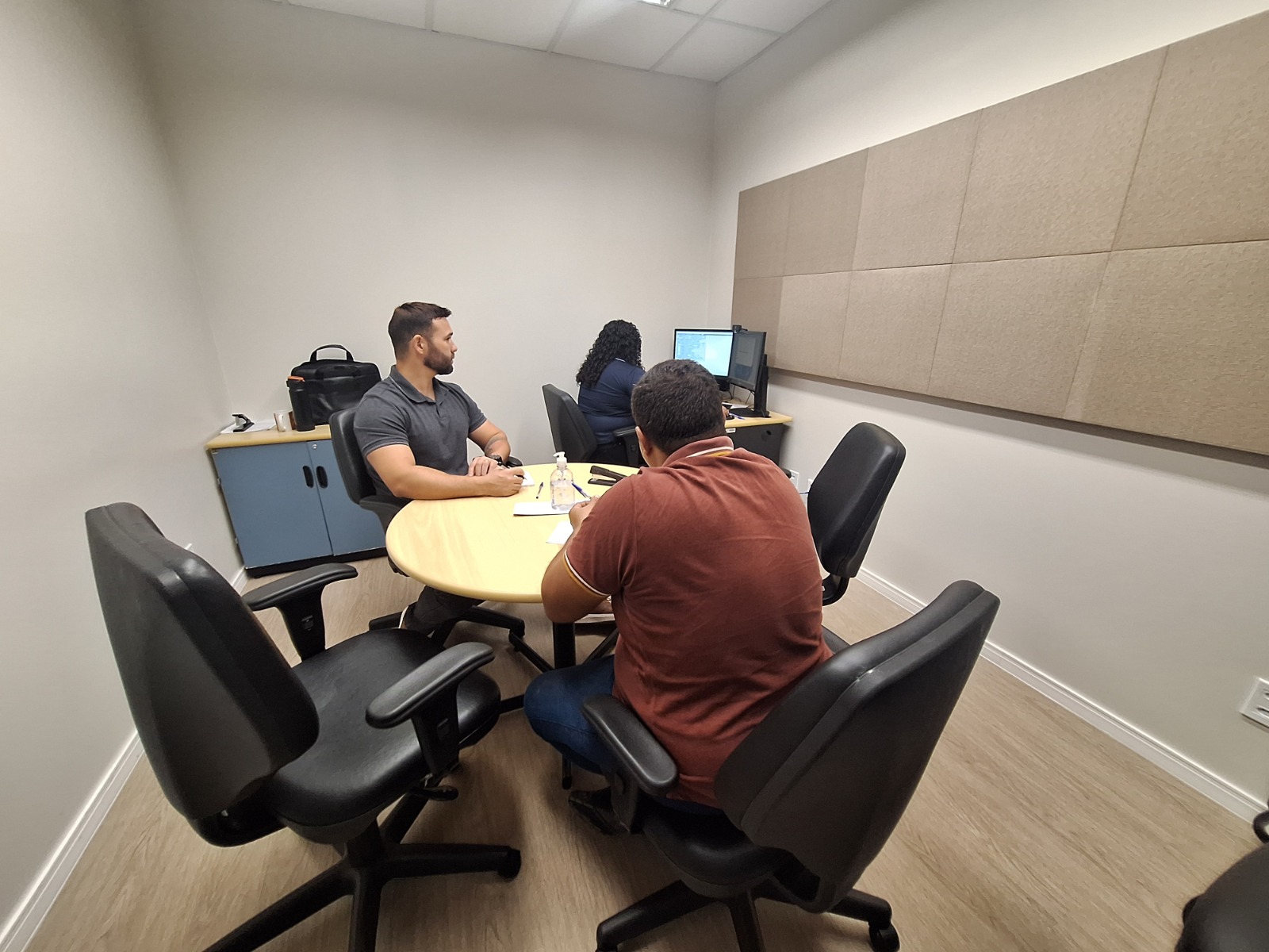 Foto 2: Em uma sala de atendimento, uma mulher de roupa rosa conversa com uma atendente de óculos e casaco azul, que sorri enquanto escreve em um papel. Ao fundo, outra pessoa trabalha em um computador. Sobre a mesa há copos, papéis e um frasco de álcool em gel. O espaço é iluminado e organizado, com cadeiras pretas e paredes claras. Foto 2: Em uma sala de atendimento, uma mulher de roupa rosa conversa com uma atendente de óculos e casaco azul, que sorri enquanto escreve em um papel. Ao fundo, outra pessoa trabalha em um computador. Sobre a mesa há copos, papéis e um frasco de álcool em gel. O espaço é iluminado e organizado, com cadeiras pretas e paredes claras.