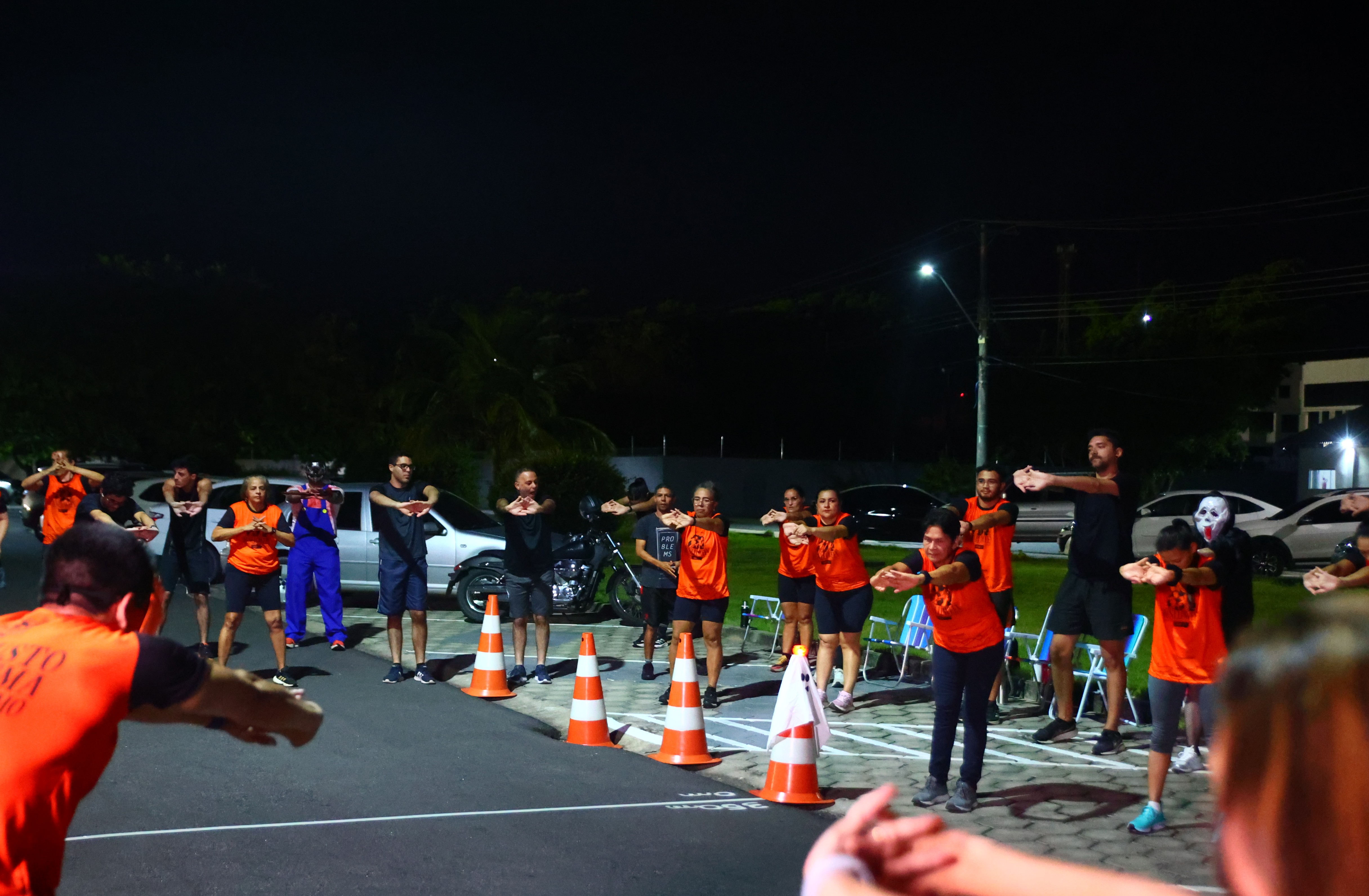 Um grupo de pessoas, muitas vestindo camisetas laranja e algumas fantasiadas, está realizando exercícios de aquecimento ou alongamento em uma rua ou estacionamento à noite. Há cones de trânsito alaranjados dispostos no chão, e a cena é iluminada por postes de luz e luzes ambientais. A pessoa em primeiro plano está de costas, também em uma camiseta laranja.