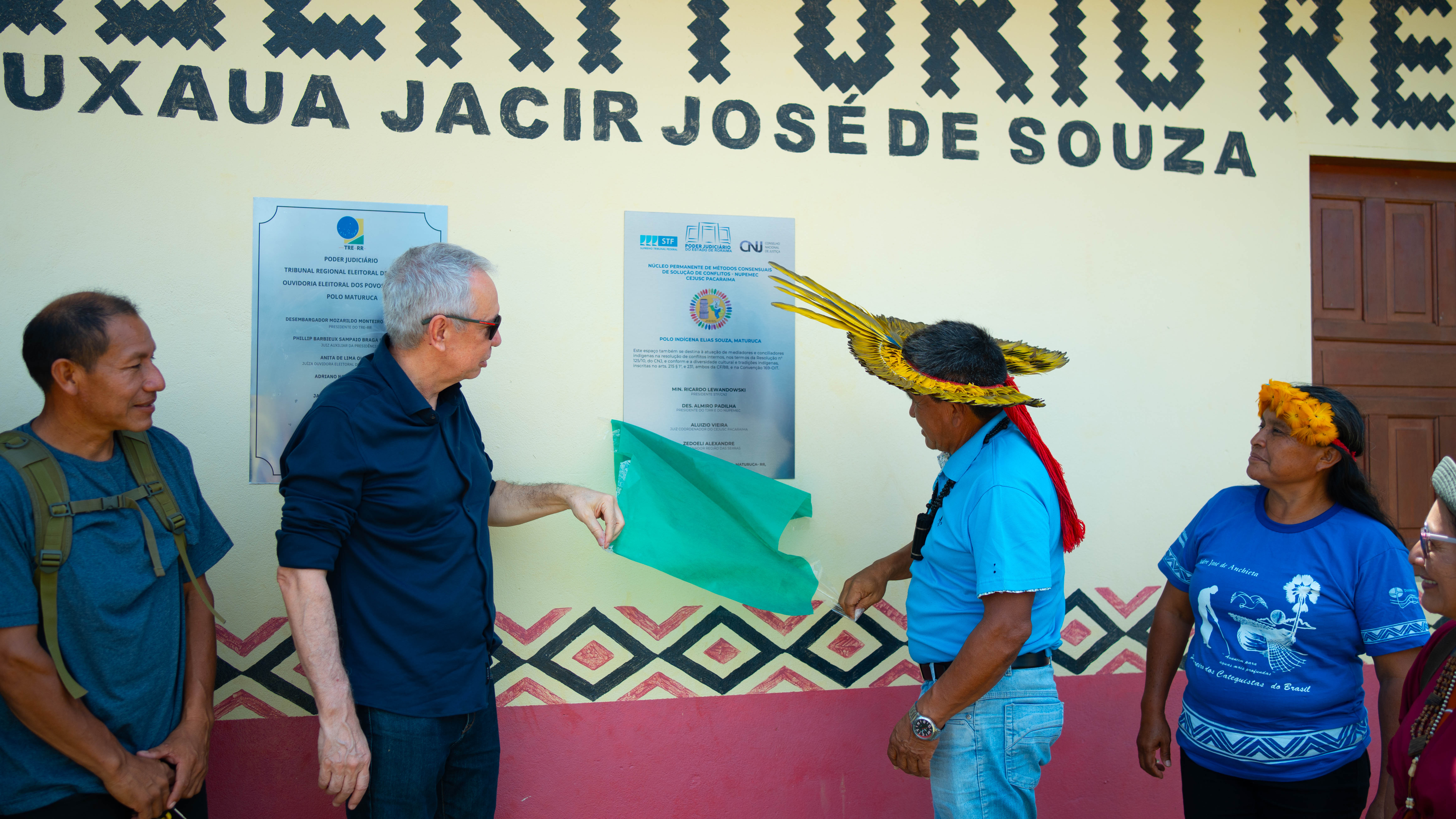 Quatro pessoas estão sentadas ao redor de uma mesa. Um homem de camisa escura aponta para uma grande placa metálica da placa de inauguração, com logotipos do STF, CNJ e CNMP. Há uma estatueta da Justiça e um cartaz da "Ouvidoria Eleitoral Indígena TRE-RR" na parede, indicando que a reunião pode estar relacionada a questões de justiça ou direitos indígenas.