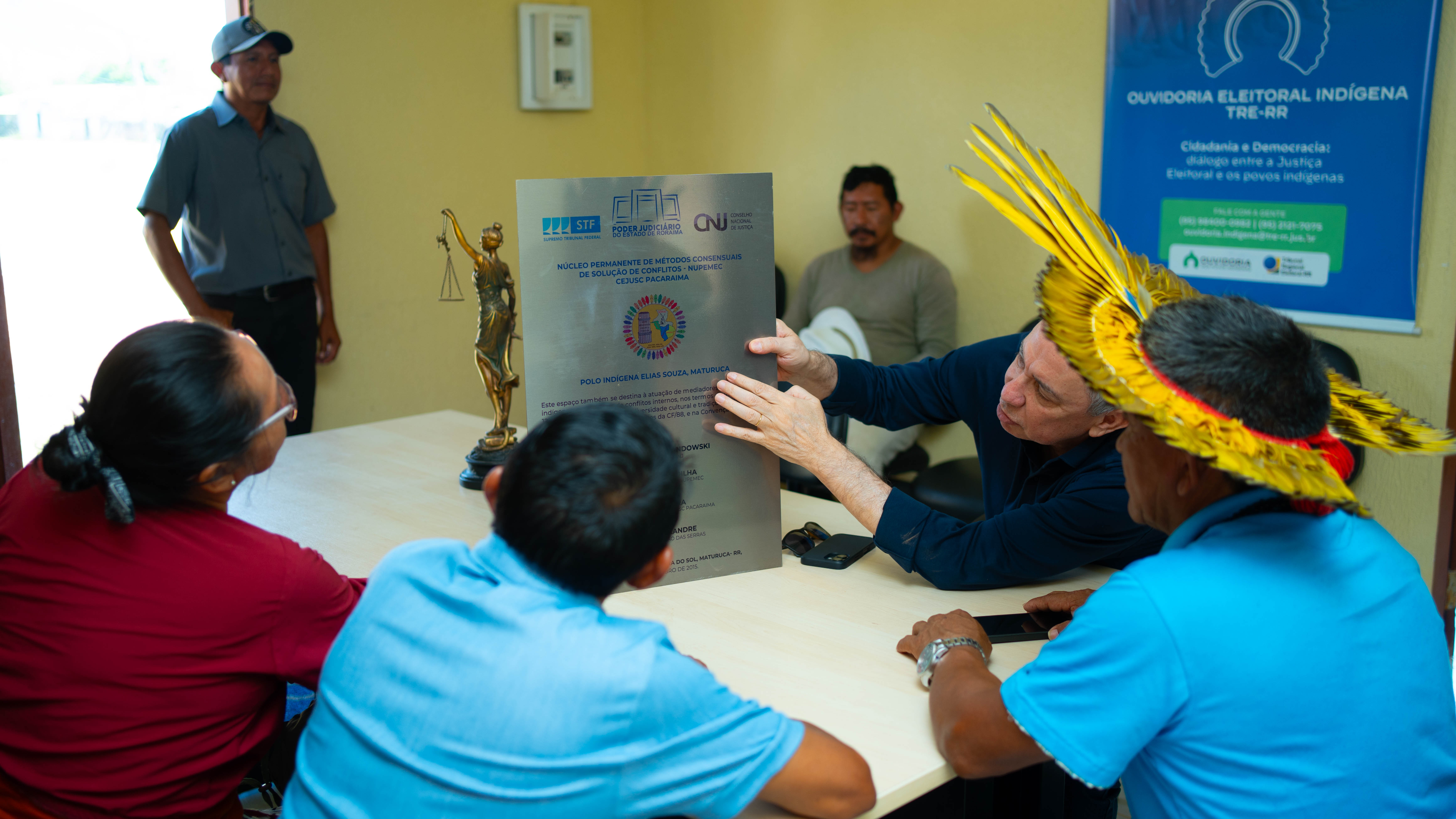 Quatro pessoas, incluindo dois homens indígenas com cocares tradicionais e um homem de camisa escura, estão em frente a uma parede de cor clara. Eles estão apontando para duas placas de inauguração fixadas na parede. A cena parece é a inauguração da revitalização no espaço