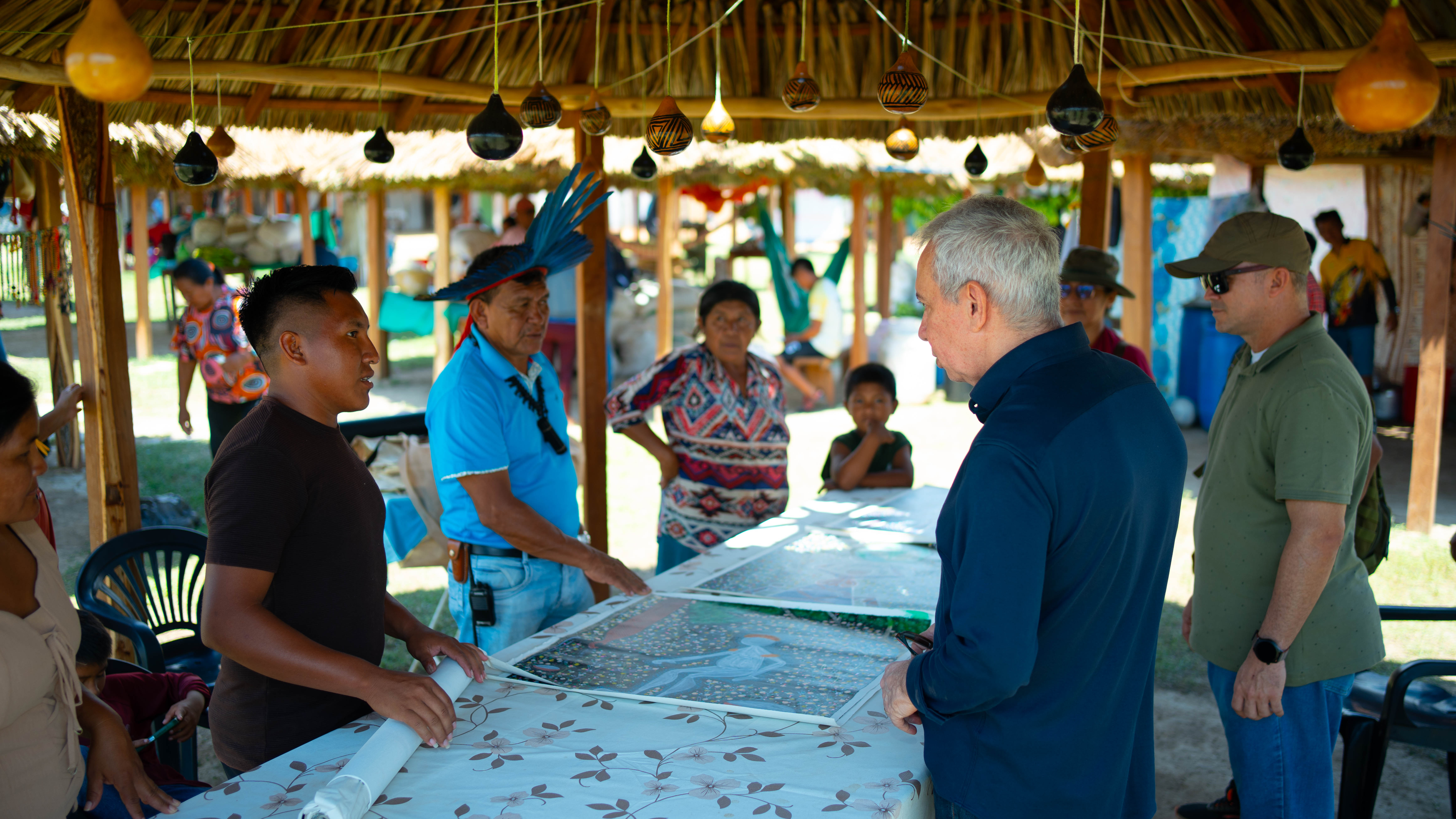 Um grupo de pessoas se reunido sob uma estrutura rústica de telhado de palha, que tem decorações penduradas. Eles estão olhando para uma grande peça de artesanato ou arte colocada sobre uma mesa durante uma feira na comunidade indígena.