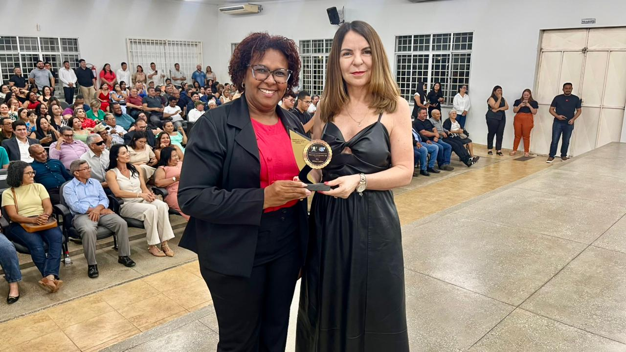 A juíza titular da Comarc Liliane Cardoso ao lado da vereadora Cleudimar Brito em um palco ou frente de sala. A mulher à esquerda, de blazer e óculos, segura um prêmio redondo dourado. Ambas sorriem para a câmera, e ao fundo, um grande público está sentado em cadeiras, assistindo a uma cerimônia ou evento. A juíza titular da Comarc Liliane Cardoso ao lado da vereadora Cleudimar Brito em um palco ou frente de sala. A mulher à esquerda, de blazer e óculos, segura um prêmio redondo dourado. Ambas sorriem para a câmera, e ao fundo, um grande público está sentado em cadeiras, assistindo a uma cerimônia ou evento.