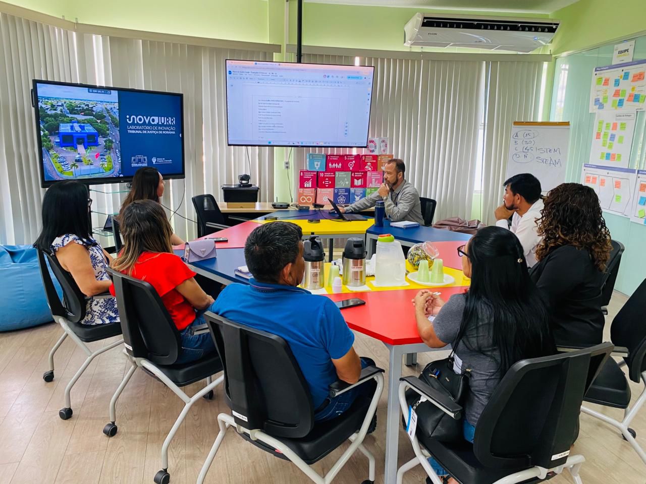 Uma reunião está acontecendo no laboratório de inovação ou sala de treinamento. Um grupo de cerca de oito pessoas está sentado em torno de uma mesa hexagonal colorida, olhando para um homem no centro que está interagindo com uma grande tela. As telas exibem um slide com o logotipo "INOVARJURR" (Laboratório de Inovação do Tribunal de Justiça de Roraima) e o outro parece ter um documento de texto ou planilha. Há cartazes com os Objetivos de Desenvolvimento Sustentável (ODS) da ONU na parede de trás, sugerindo um foco em inovação e planejamento. Uma reunião está acontecendo no laboratório de inovação ou sala de treinamento. Um grupo de cerca de oito pessoas está sentado em torno de uma mesa hexagonal colorida, olhando para um homem no centro que está interagindo com uma grande tela. As telas exibem um slide com o logotipo "INOVARJURR" (Laboratório de Inovação do Tribunal de Justiça de Roraima) e o outro parece ter um documento de texto ou planilha. Há cartazes com os Objetivos de Desenvolvimento Sustentável (ODS) da ONU na parede de trás, sugerindo um foco em inovação e planejamento.