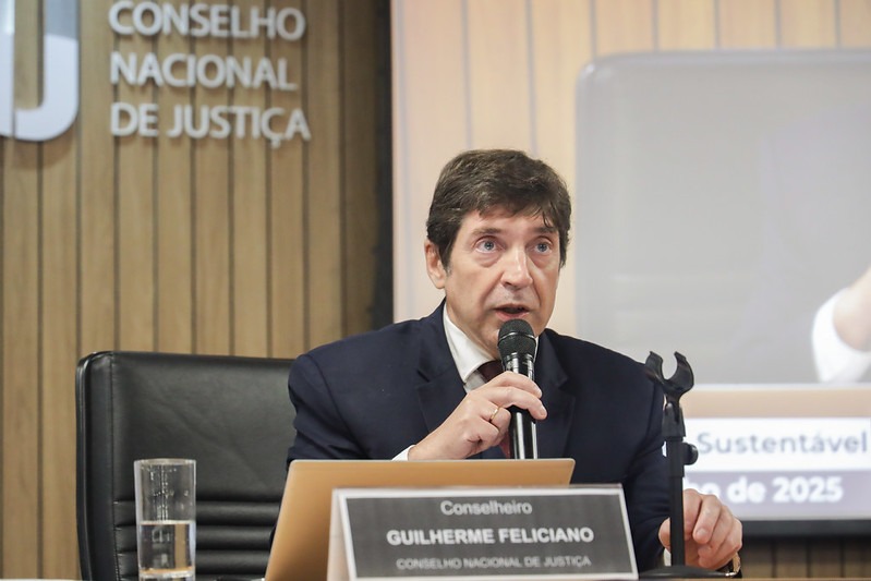  A imagem mostra o conselheiro Guilherme Guimarães Feliciano, de terno azul-marinho e camisa branca, falando ao microfone durante uma reunião. Ele está sentado à mesa, em frente a um notebook dourado, com uma placa de identificação à sua frente onde se lê “Conselheiro Guilherme Feliciano – Conselho Nacional de Justiça”. Ao fundo, há o logotipo do CNJ (Conselho Nacional de Justiça) em uma parede de madeira clara. Um copo com água está sobre a mesa ao lado esquerdo. A cena ocorre em um ambiente institucional, com iluminação neutra.