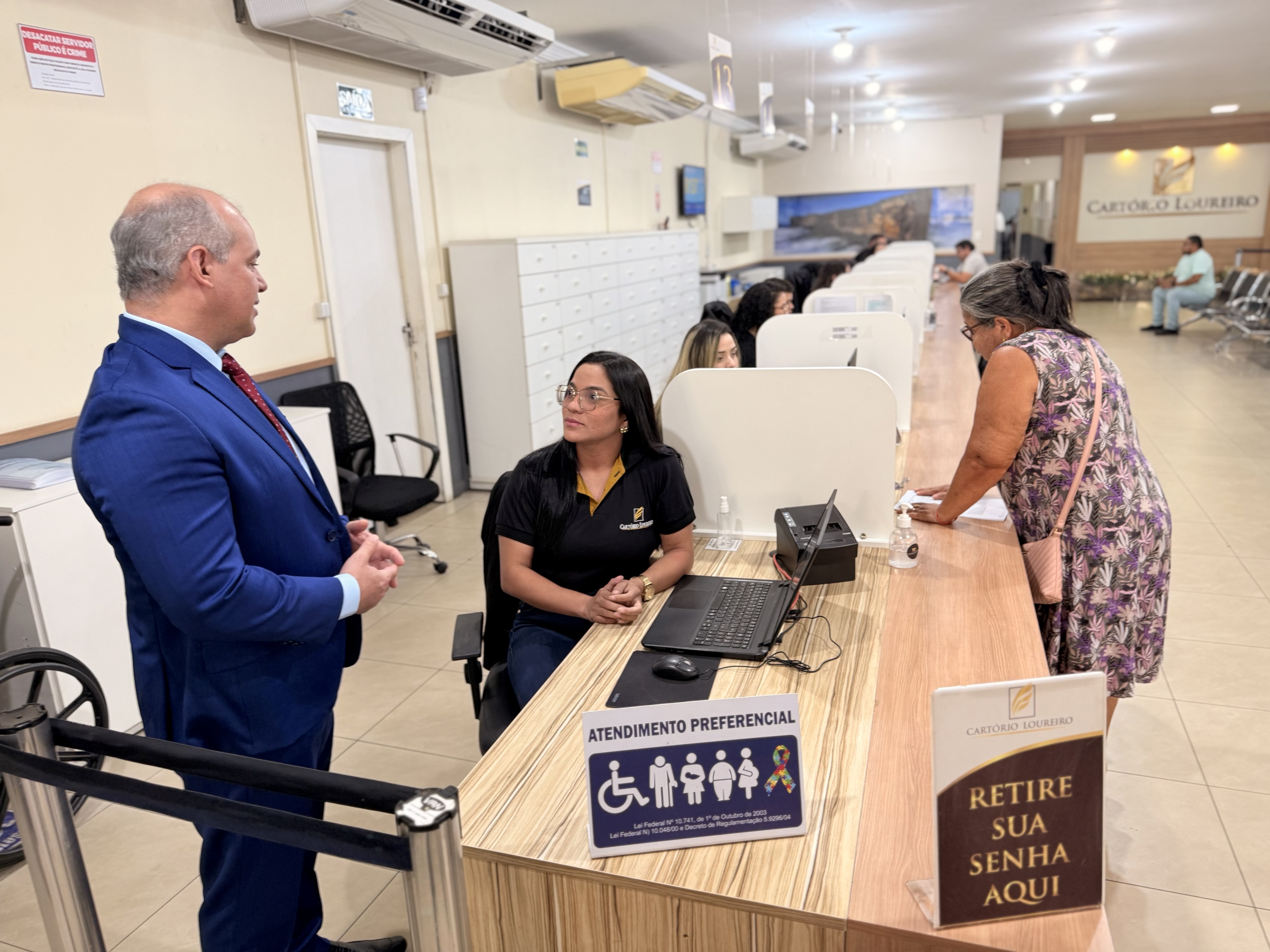 Imagem colorida mostra a vista de um guichê de "Atendimento Preferencial" em um cartório. Uma atendente sentada, vestindo uma camisa preta com logo, conversa com o juiz-auxiliar da CGJ/TJRR. Uma mulher idosa, com óculos e vestindo um vestido florido, aproxima-se do guichê pela direita. Sobre o balcão de madeira, há um aviso de "Atendimento Preferencial" e uma placa para "Retire sua senha aqui" com o logo "Cartório Loureiro". Ao fundo, é possível ver outros guichês e uma área de espera. Imagem colorida mostra a vista de um guichê de "Atendimento Preferencial" em um cartório. Uma atendente sentada, vestindo uma camisa preta com logo, conversa com o juiz-auxiliar da CGJ/TJRR. Uma mulher idosa, com óculos e vestindo um vestido florido, aproxima-se do guichê pela direita. Sobre o balcão de madeira, há um aviso de "Atendimento Preferencial" e uma placa para "Retire sua senha aqui" com o logo "Cartório Loureiro". Ao fundo, é possível ver outros guichês e uma área de espera.