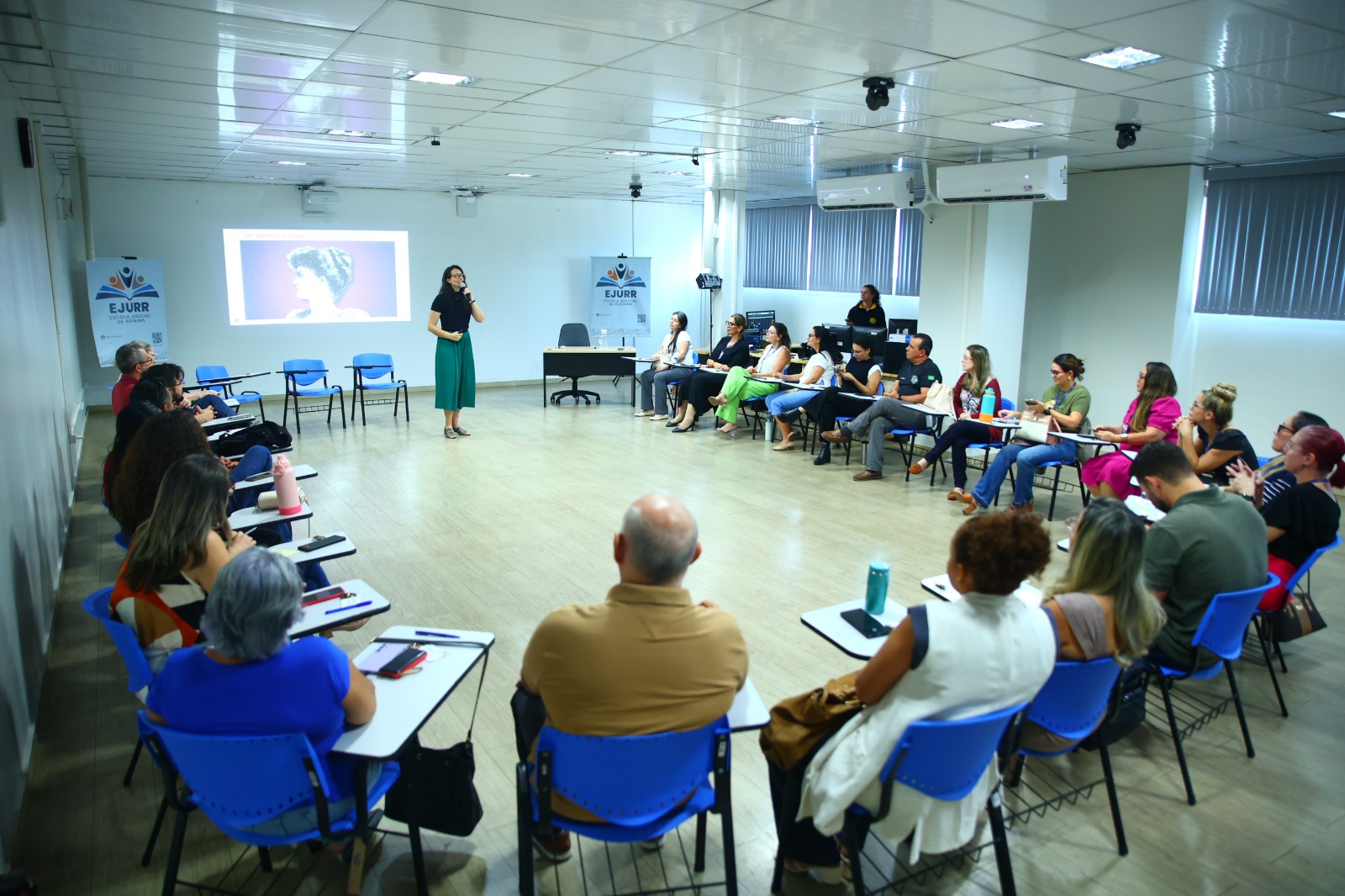 Uma sala com participantes em um workshop, onde uma apresentadora fala para o grupo. Há cadeiras dispostas em semicírculo e um projetor exibindo uma imagem na parede.