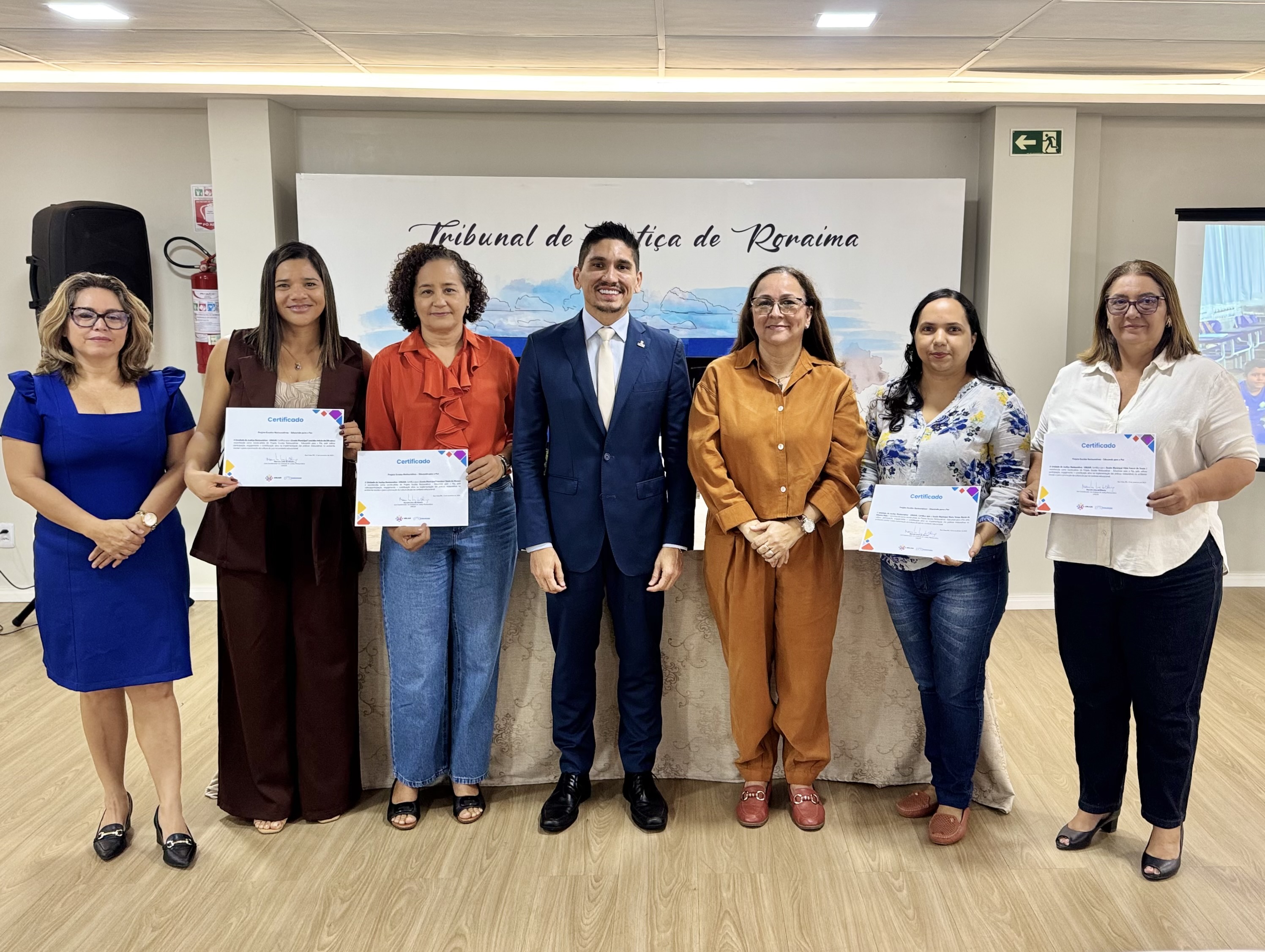Imagem colorida mostra sete pessoas em pé, lado a lado, em um ambiente interno. Três delas seguram certificados nas mãos. Ao fundo, há um painel com a inscrição "Tribunal de Justiça de Roraima" e paisagem ilustrada. Todas estão vestidas formalmente, com roupas sociais. Imagem colorida mostra sete pessoas em pé, lado a lado, em um ambiente interno. Três delas seguram certificados nas mãos. Ao fundo, há um painel com a inscrição "Tribunal de Justiça de Roraima" e paisagem ilustrada. Todas estão vestidas formalmente, com roupas sociais.
