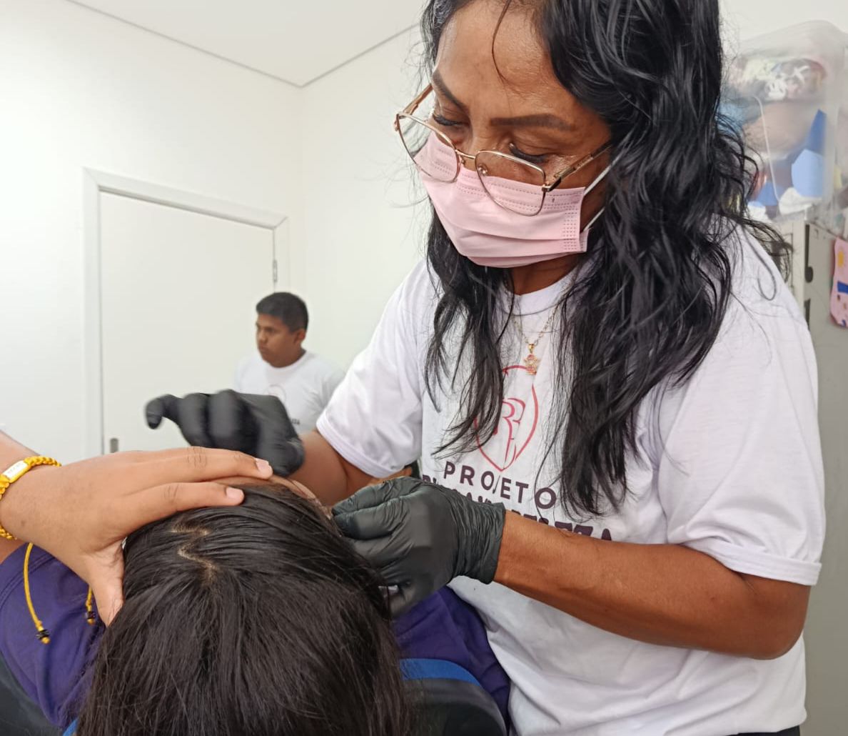 Imagem colorida mostra uma mulher usando luvas pretas manipulando o cabelo de outra pessoa que está sentada à sua frente. A mulher veste uma camiseta branca estampada com o texto "PROJETO" e parte de um logo visível. Ao fundo, aparece um homem desfocado, também de camiseta branca, e detalhes do ambiente, como uma porta branca e prateleiras. Imagem colorida mostra uma mulher usando luvas pretas manipulando o cabelo de outra pessoa que está sentada à sua frente. A mulher veste uma camiseta branca estampada com o texto "PROJETO" e parte de um logo visível. Ao fundo, aparece um homem desfocado, também de camiseta branca, e detalhes do ambiente, como uma porta branca e prateleiras.