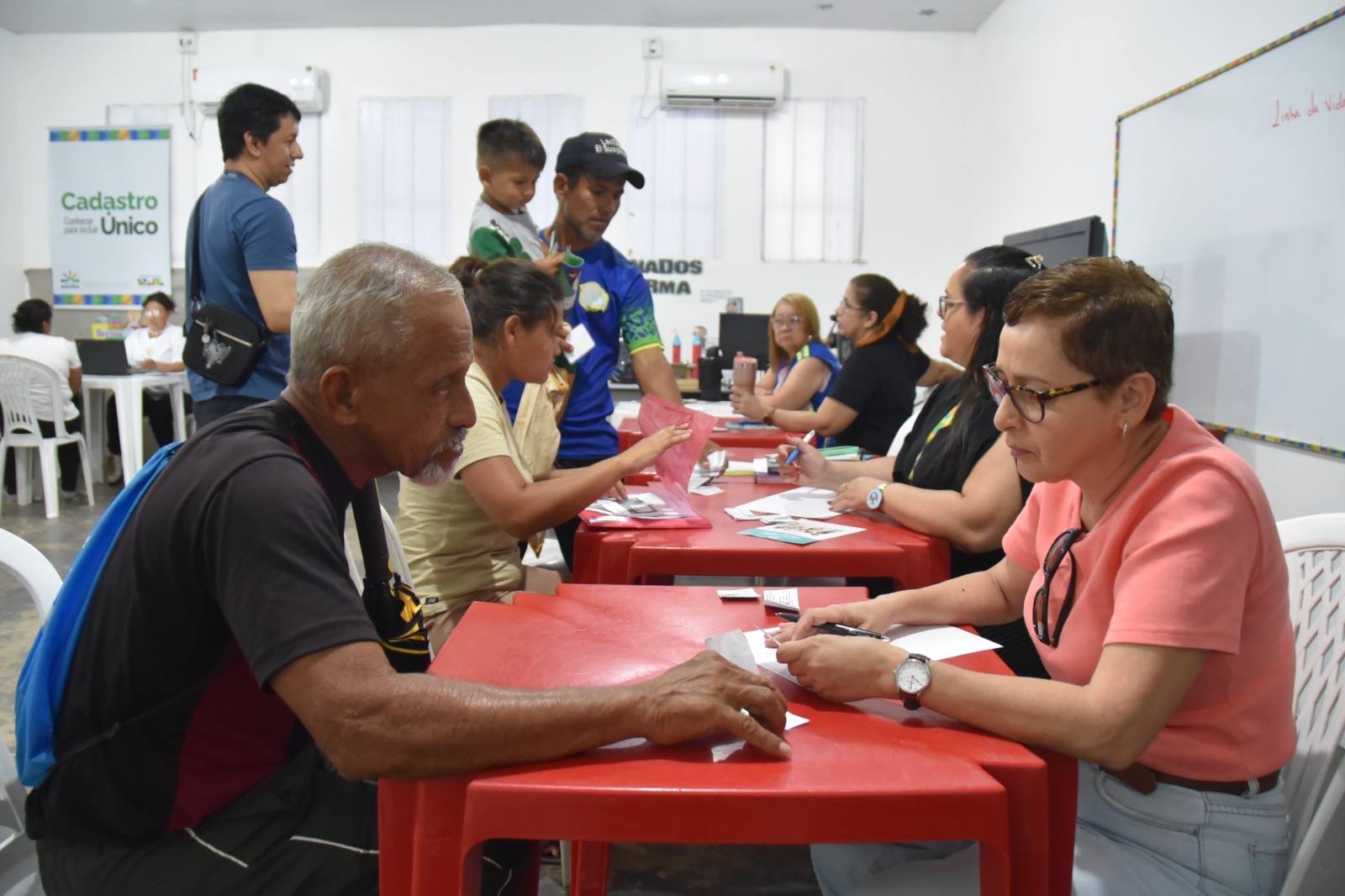Imagem de um evento de cadastro, com pessoas sentadas em mesas vermelhas se consultando. Um homem idoso interage com uma mulher, enquanto outras pessoas, incluindo um menino e trabalhadores, estão ao fundo, em um ambiente bem iluminado e organizado. Imagem de um evento de cadastro, com pessoas sentadas em mesas vermelhas se consultando. Um homem idoso interage com uma mulher, enquanto outras pessoas, incluindo um menino e trabalhadores, estão ao fundo, em um ambiente bem iluminado e organizado.