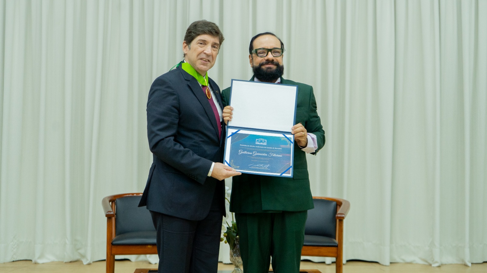 Conselheiro Guilherme Guimarães Feliciano ao lado do presidente do TJRR, desembargador Leonardo Cupello, após ser agraciado com a Outorga da Medalha do Mérito Judiciário do Estado de Roraima. Ambos seguram um certificado. O homem à esquerda usa um terno escuro e uma gravata, enquanto o da direita veste um terno verde. Ao fundo, há cortinas brancas e cadeiras de madeira.