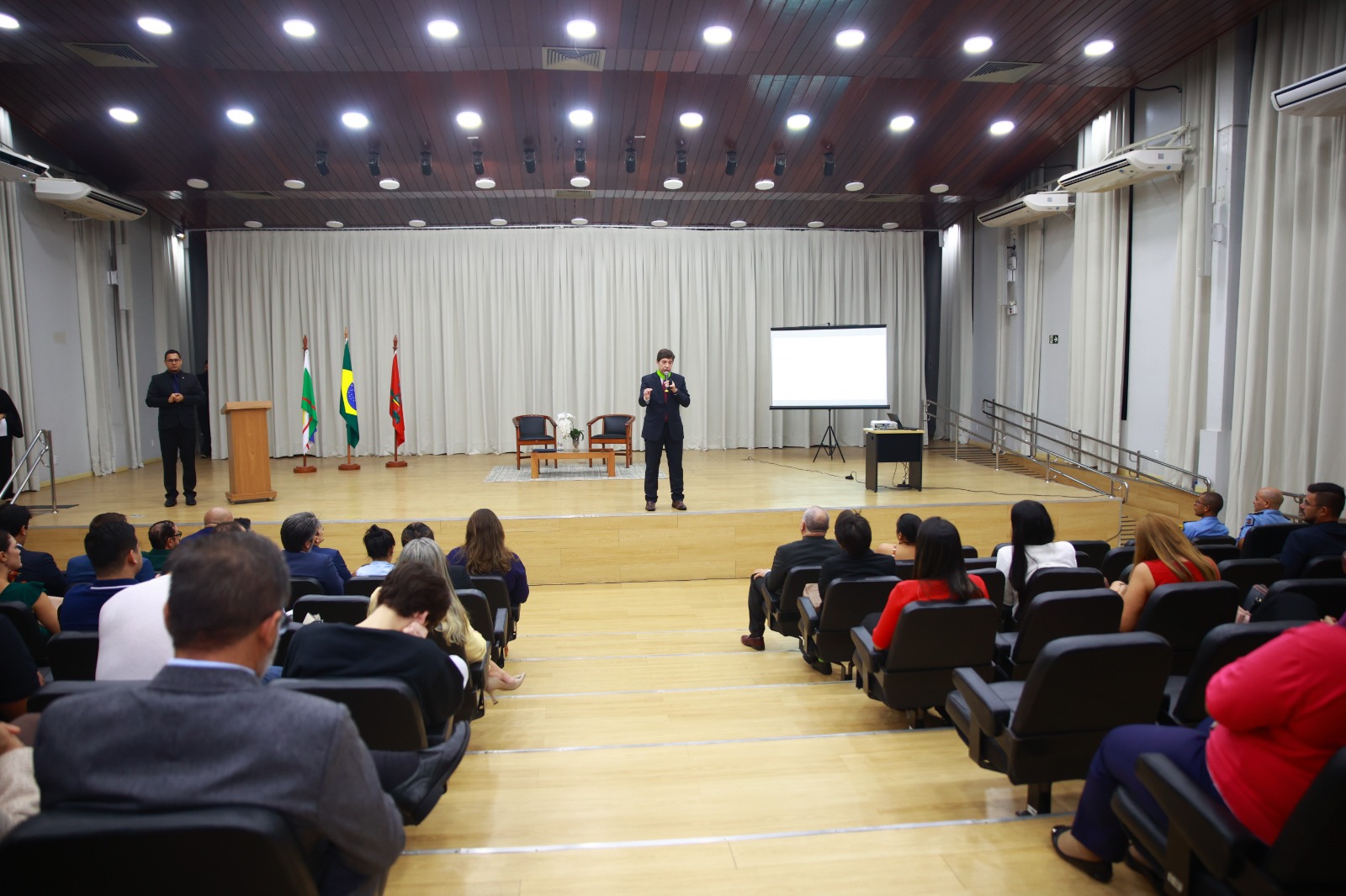 Foto colorida mostra o público em uma sala de conferência, assistindo a apresentação do conselheiro Guilherme Guimarães. 