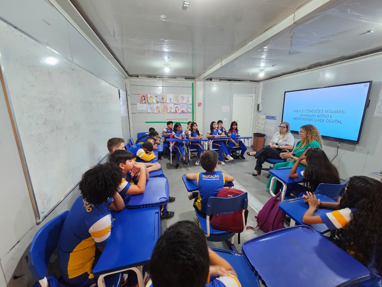 A imagem mostra uma sala de aula com várias crianças sentadas em carteiras azuis, todas usando uniforme escolar. Duas mulheres conversando com a turma.