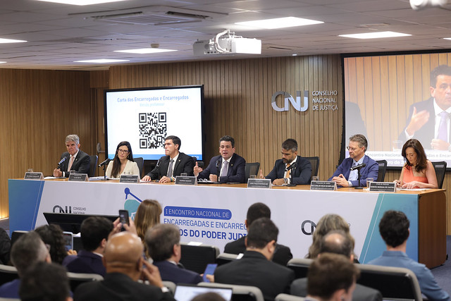 foto colorida mostra um painel de debate no Conselho Nacional de Justiça (CNJ). Sete participantes (cinco homens e duas mulheres) estão sentados em uma mesa, de frente para uma plateia. Um banner na frente da mesa identifica o evento como o "I Encontro Nacional de Encarregadas e Encarregados do Poder Judiciário". Ao fundo, uma tela exibe o título "Carta dos Encarregados e Encarregadas" com um QR Code. foto colorida mostra um painel de debate no Conselho Nacional de Justiça (CNJ). Sete participantes (cinco homens e duas mulheres) estão sentados em uma mesa, de frente para uma plateia. Um banner na frente da mesa identifica o evento como o "I Encontro Nacional de Encarregadas e Encarregados do Poder Judiciário". Ao fundo, uma tela exibe o título "Carta dos Encarregados e Encarregadas" com um QR Code.