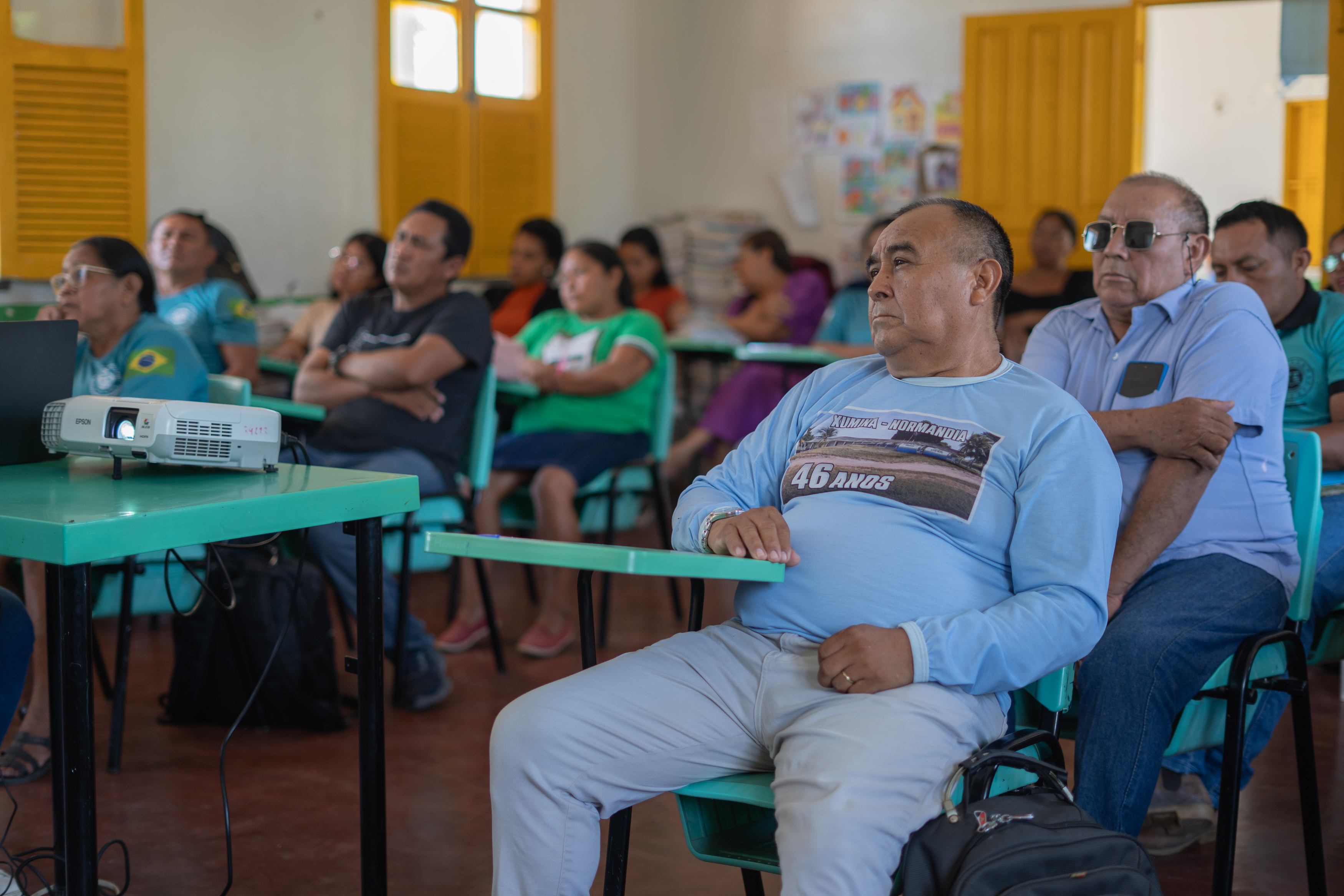 Imagem de uma sala de aula com várias pessoas assistindo a uma apresentação. Um projetor está colocado na mesa à frente, e os participantes, em sua maioria sentados, mostram expressões de atenção. O ambiente tem paredes claras e janelas com persianas amarelas. Imagem de uma sala de aula com várias pessoas assistindo a uma apresentação. Um projetor está colocado na mesa à frente, e os participantes, em sua maioria sentados, mostram expressões de atenção. O ambiente tem paredes claras e janelas com persianas amarelas.