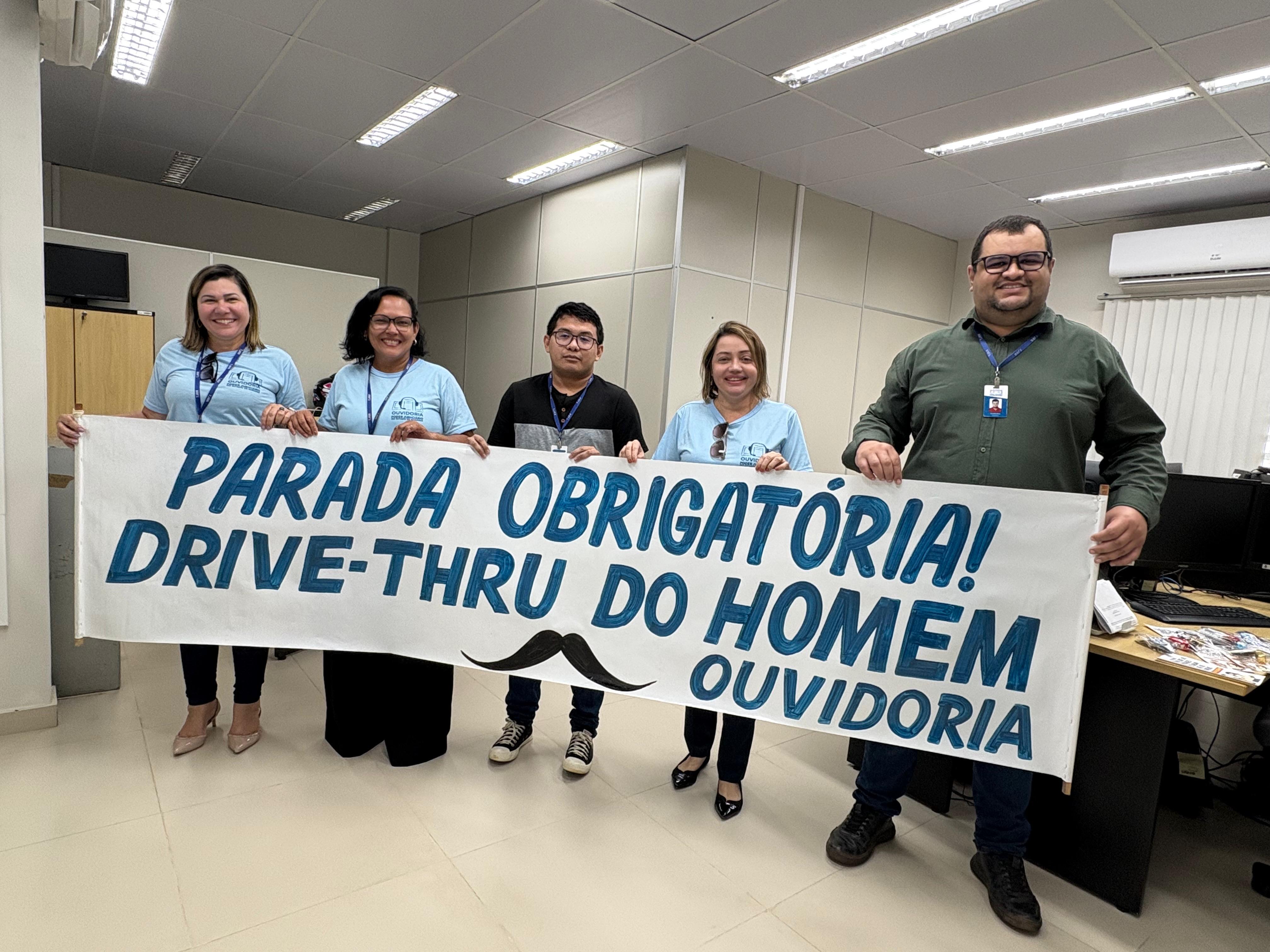  imagem colorida mostra 5 pessoas, sendo 2 mulheres e 3 homens segurando uma faixa com o texto "Parada obrigatória Drive thru do homem ouvidoria". 