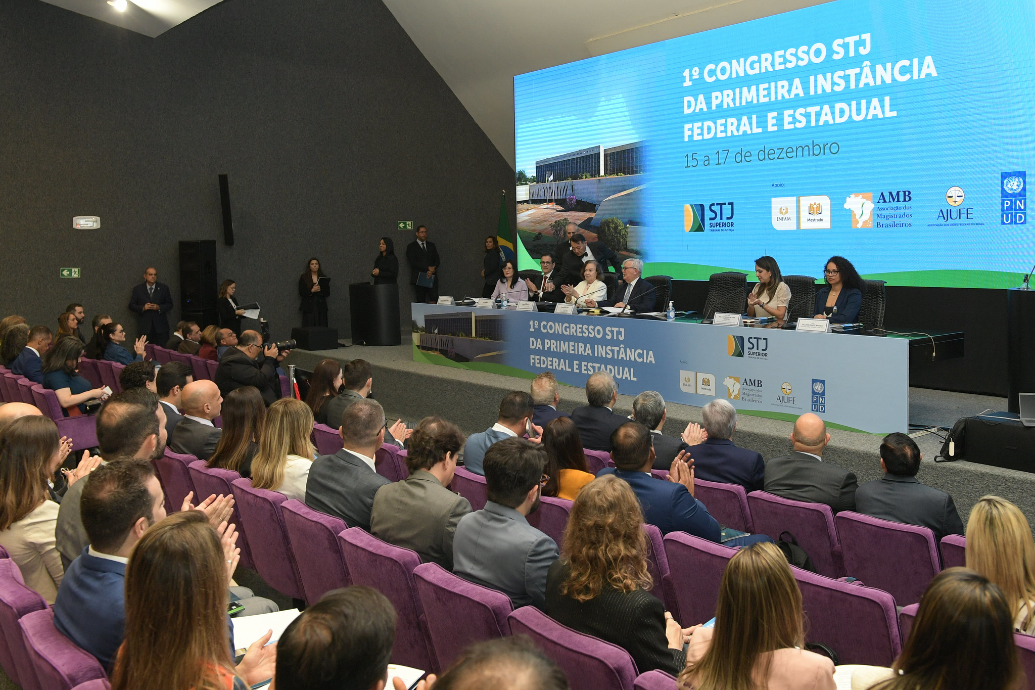 Imagem colorida contém um auditório lotado durante um congresso. No palco, autoridades estão sentadas em uma longa mesa branca. Ao fundo, um telão exibe o título: "1º Congresso STJ da Primeira Instância Federal e Estadual". A plateia, majoritariamente em trajes sociais, está sentada em cadeiras roxas, aplaudindo o painel. Imagem colorida contém um auditório lotado durante um congresso. No palco, autoridades estão sentadas em uma longa mesa branca. Ao fundo, um telão exibe o título: "1º Congresso STJ da Primeira Instância Federal e Estadual". A plateia, majoritariamente em trajes sociais, está sentada em cadeiras roxas, aplaudindo o painel.