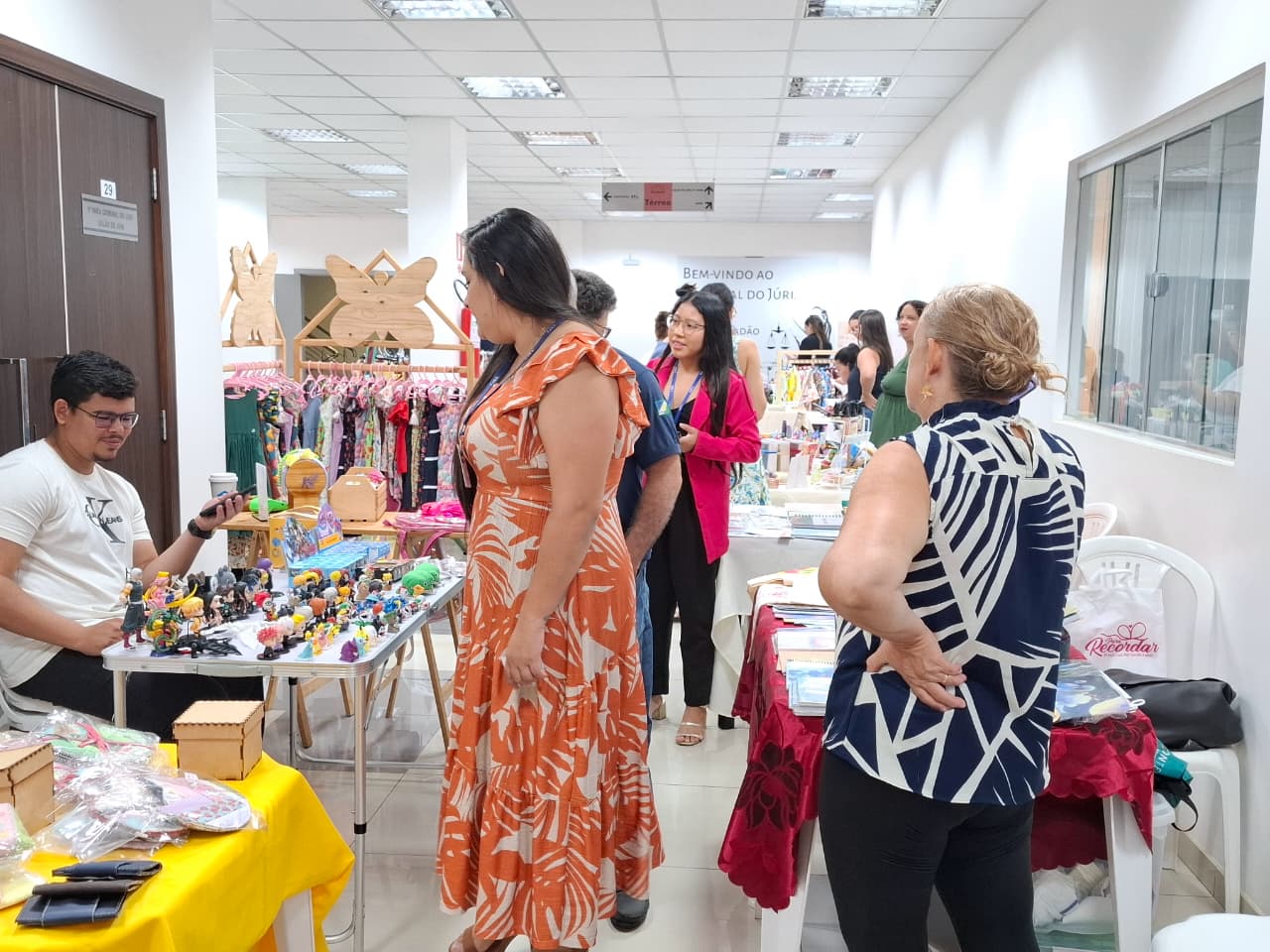 Imagem colorida mostra uma feira de artesanato acontecendo em um corredor. Várias pessoas estão em frente a mesas com produtos expostos. Em primeiro plano, à esquerda, um homem com camisa branca está sentado atrás de uma mesa, interagindo com uma mulher que usa um vestido estampado. Outras pessoas estão espalhadas pelo fundo do corredor, olhando os produtos.