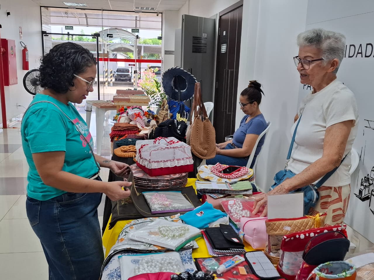  Imagem colorida mostra uma feira de artesanato. Em primeiro plano, uma mulher com uma blusa turquesa está examinando os produtos em uma mesa cheia de itens, enquanto uma mulher mais velha de cabelos brancos conversa com ela. Os produtos incluem bolsas, chapéus, e outros acessórios feitos à mão.