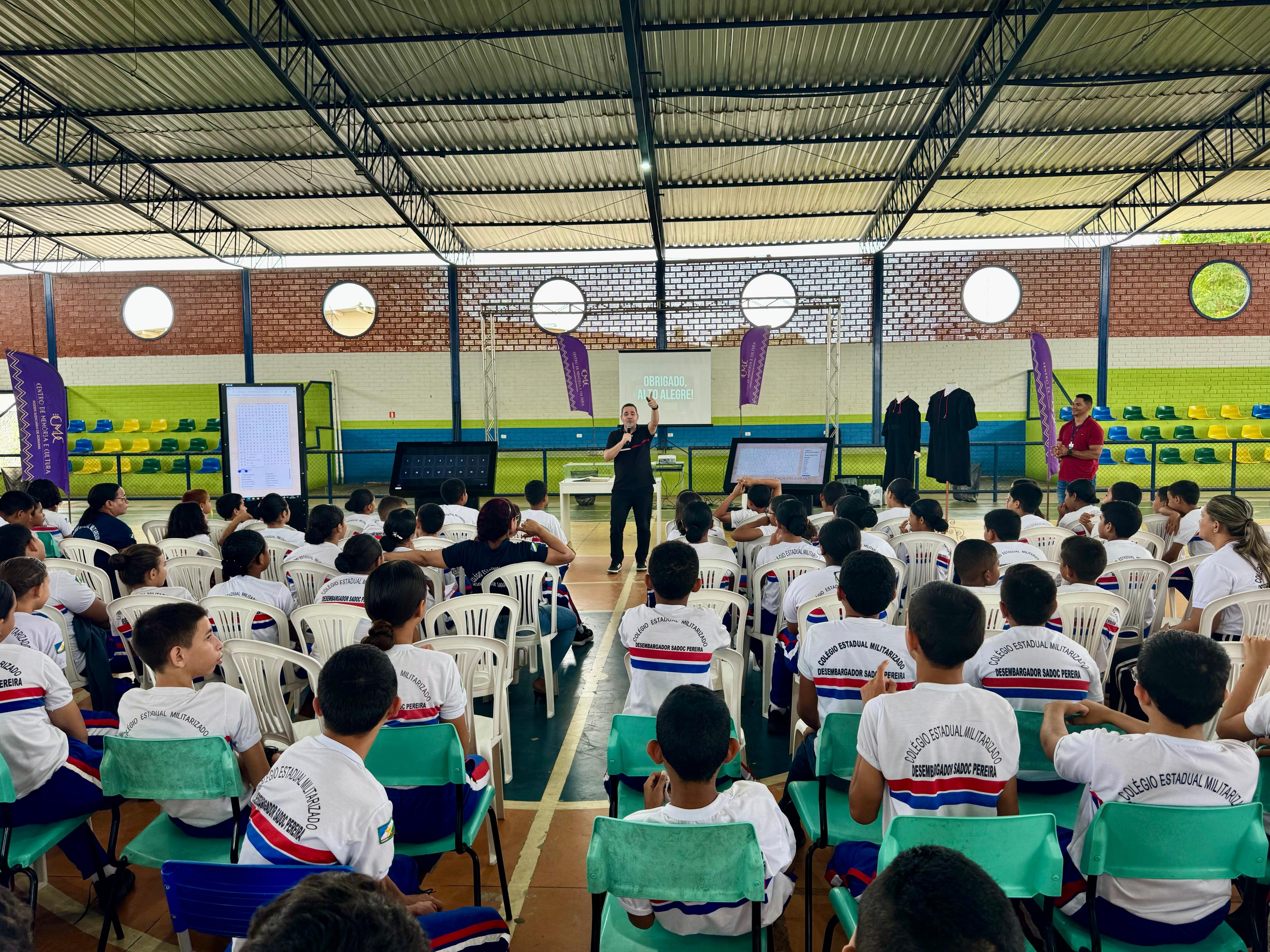 A imagem colorida mostra um grupo de jovens, a maioria vestindo camisetas brancas com logotipos coloridos, sentados em cadeiras brancas e verdes, de costas para a câmera. Eles estão em um espaço amplo, que parece ser um ginásio ou auditório, com teto alto e vigas metálicas. À frente da plateia, um homem em pé, no centro, gesticula enquanto se dirige ao público. Ao seu lado, há telões e, à direita, algumas pessoas em pé e cadeiras vazias. A imagem colorida mostra um grupo de jovens, a maioria vestindo camisetas brancas com logotipos coloridos, sentados em cadeiras brancas e verdes, de costas para a câmera. Eles estão em um espaço amplo, que parece ser um ginásio ou auditório, com teto alto e vigas metálicas. À frente da plateia, um homem em pé, no centro, gesticula enquanto se dirige ao público. Ao seu lado, há telões e, à direita, algumas pessoas em pé e cadeiras vazias.