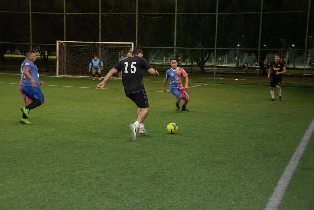 Imagem colorida mostra um momento de jogo em um campo de futebol sintético. Um jogador de costas, usando uma camisa preta com o número "15", está no centro da ação, chutando uma bola amarela. Outros jogadores, um de uniforme azul e outro de preto, também estão em movimento no campo. Ao fundo, pode-se ver um gol com rede branca e um goleiro com uniforme azul. A cena é iluminada artificialmente, sugerindo que o jogo ocorre ao anoitecer ou em um ambiente coberto. Imagem colorida mostra um momento de jogo em um campo de futebol sintético. Um jogador de costas, usando uma camisa preta com o número "15", está no centro da ação, chutando uma bola amarela. Outros jogadores, um de uniforme azul e outro de preto, também estão em movimento no campo. Ao fundo, pode-se ver um gol com rede branca e um goleiro com uniforme azul. A cena é iluminada artificialmente, sugerindo que o jogo ocorre ao anoitecer ou em um ambiente coberto.
