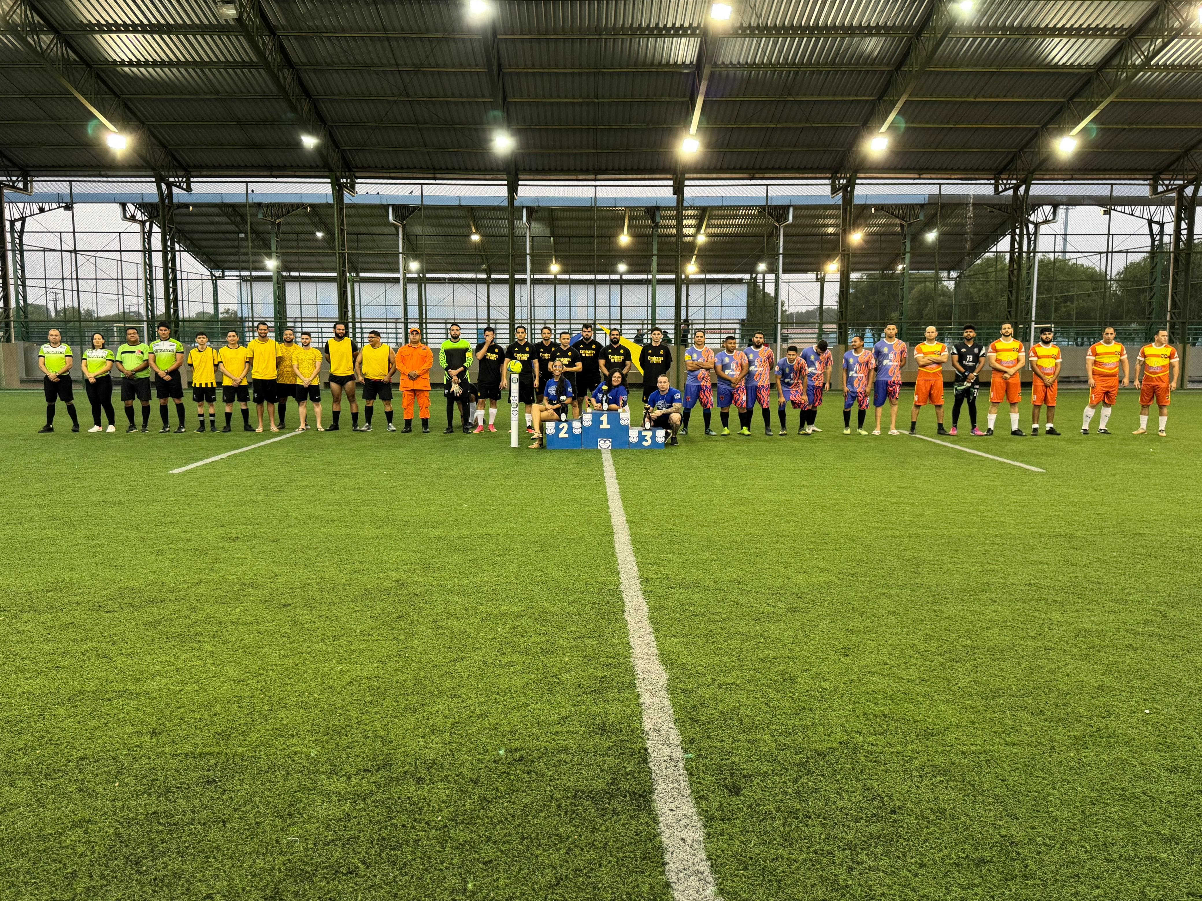 Imagem colorida mostra uma vista ampla de um torneio de futebol, com diversos times alinhados no centro de um campo de grama sintética coberto. Os jogadores vestem uniformes de cores variadas, incluindo preto, amarelo, laranja, azul e um padrão colorido. Na frente dos times, estão dispostos três pódios numerados (1, 2 e 3). O teto da quadra e a iluminação artificial estão visíveis. Imagem colorida mostra uma vista ampla de um torneio de futebol, com diversos times alinhados no centro de um campo de grama sintética coberto. Os jogadores vestem uniformes de cores variadas, incluindo preto, amarelo, laranja, azul e um padrão colorido. Na frente dos times, estão dispostos três pódios numerados (1, 2 e 3). O teto da quadra e a iluminação artificial estão visíveis.