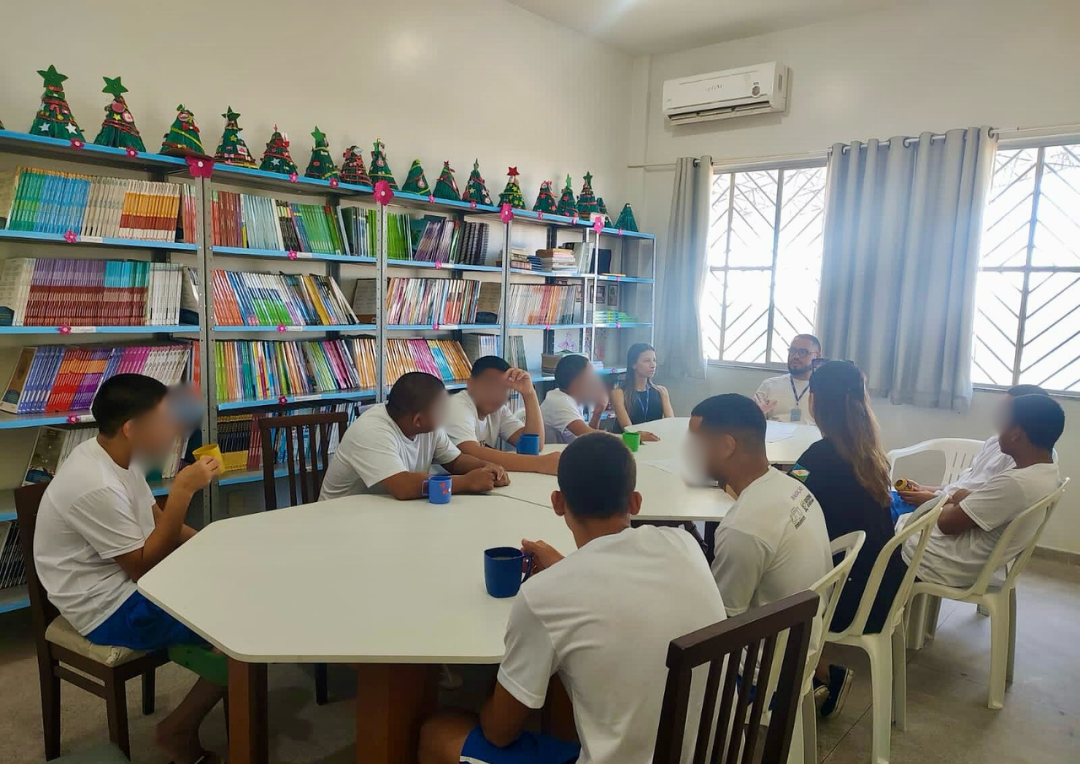 Foto colorida mostra onze pessoas, entre 2 servidores do TJRR, uma agente do GMF e oito adolescentes homens sentados em volta de uma mesa conversando. Atrás deles encontra-se uma estante de livros.