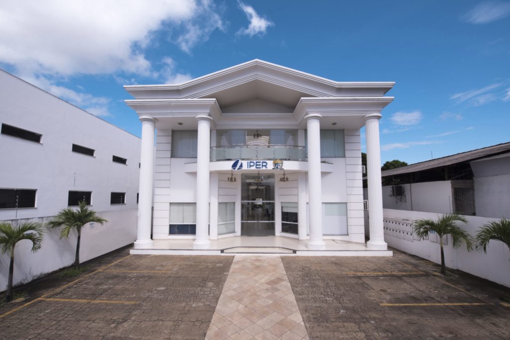 Imagem colorida mostra a fachada do prédio do Instituto de Previdência do Estado de Roraima, o edifício pintado na cor branca está identificado com o nome “IPER” pintado de Azul. Nos lados as insígnias do instituto de Previdência do Estado de Roraima e do Governo do Estado de Roraima.