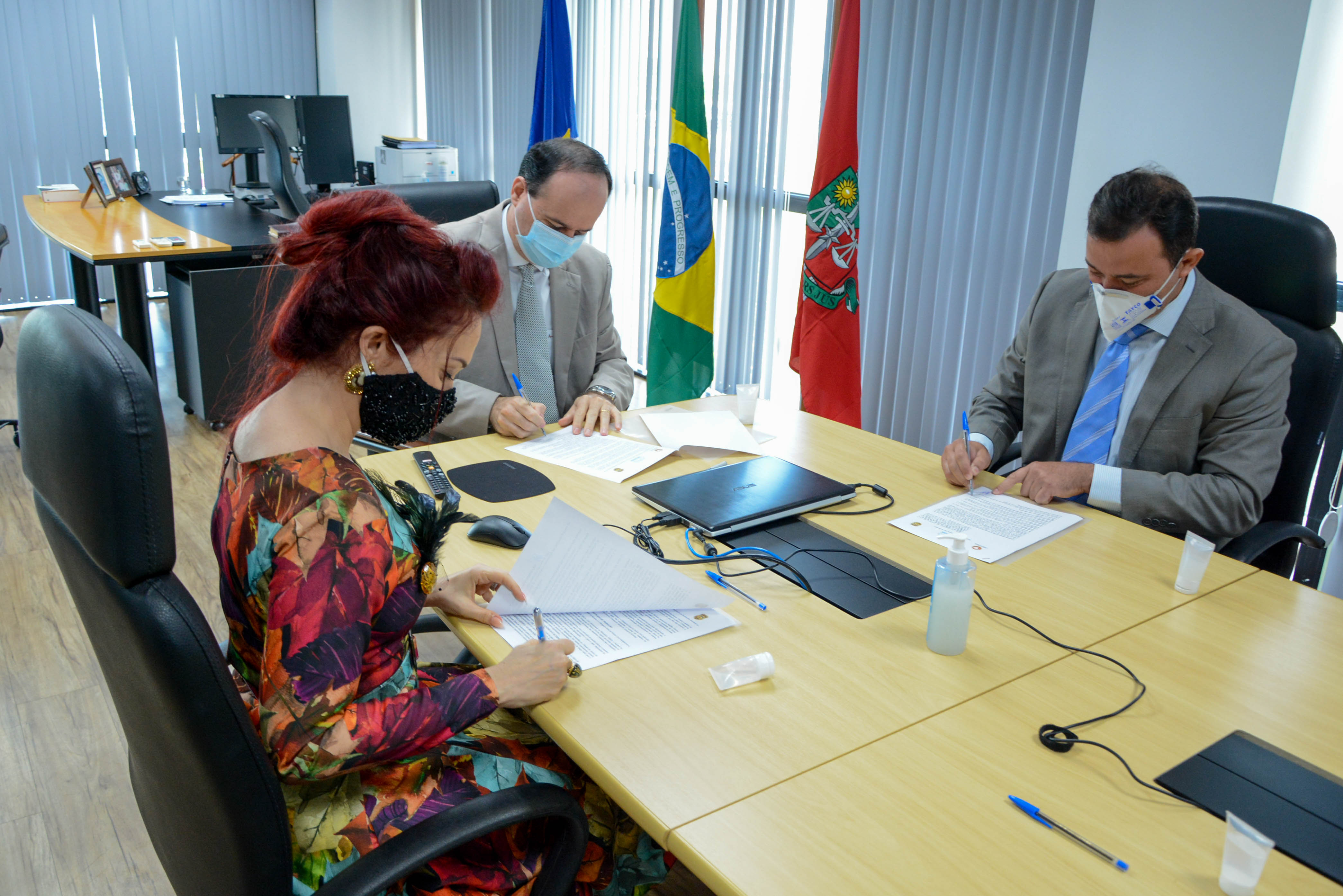 Procuradora-geral de Justiça do Ministério Público, Janaína Carneiro Costa, Delegado-geral de Polícia Civil do Estado de Roraima, Herbert de Amorim Cardoso e Presidente do Tribunal de Justiça, desembargador Cristóvão Suter, assinando documentos sentados em volta de uma mesa de reuniões. 