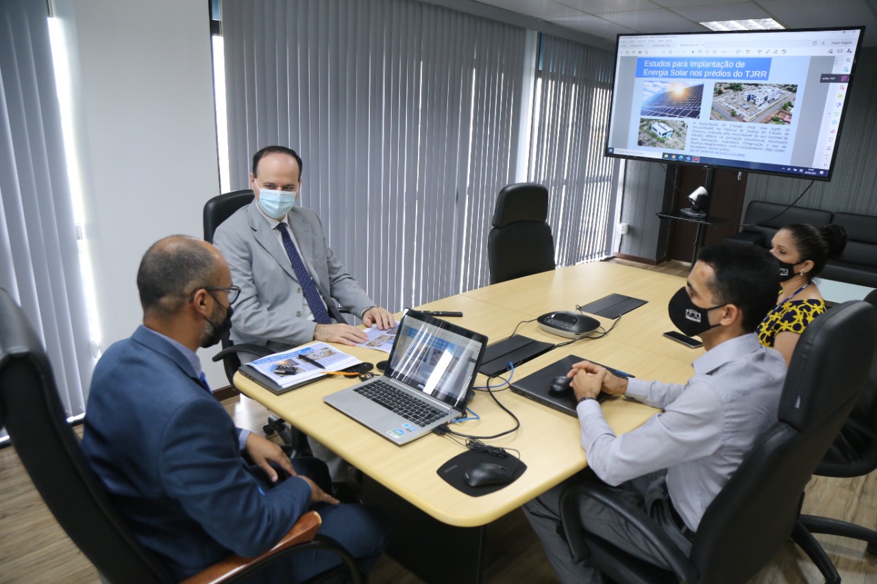 Presidente do Tribunal de Justiça, desembargador Cristóvão Suter sentado em mesa de reuniões com servidores, com um notebook sobre a mesa e uma TV ao fundo.