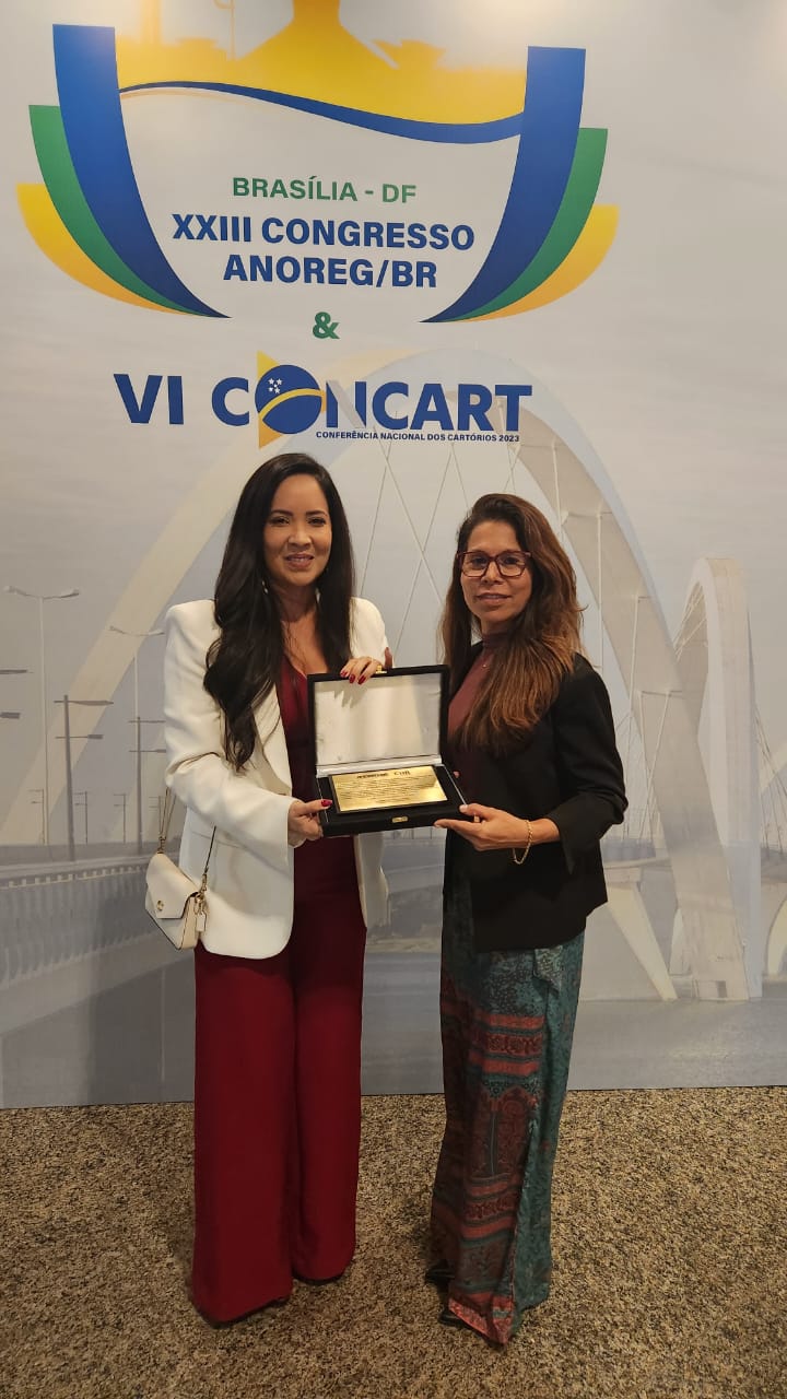  Foto colorida mostra a servidora do TJRR, Luciana Menezes,  recebendo premiação em homenagem ao desembargador Mozarildo Cavalcanti, durante congresso nacional Anoreg/BR e posando para foto ao lado da presidente da Associação dos Notários e Registradores do Estado de Roraima (ANOREG/RR), Inês Maraschin.