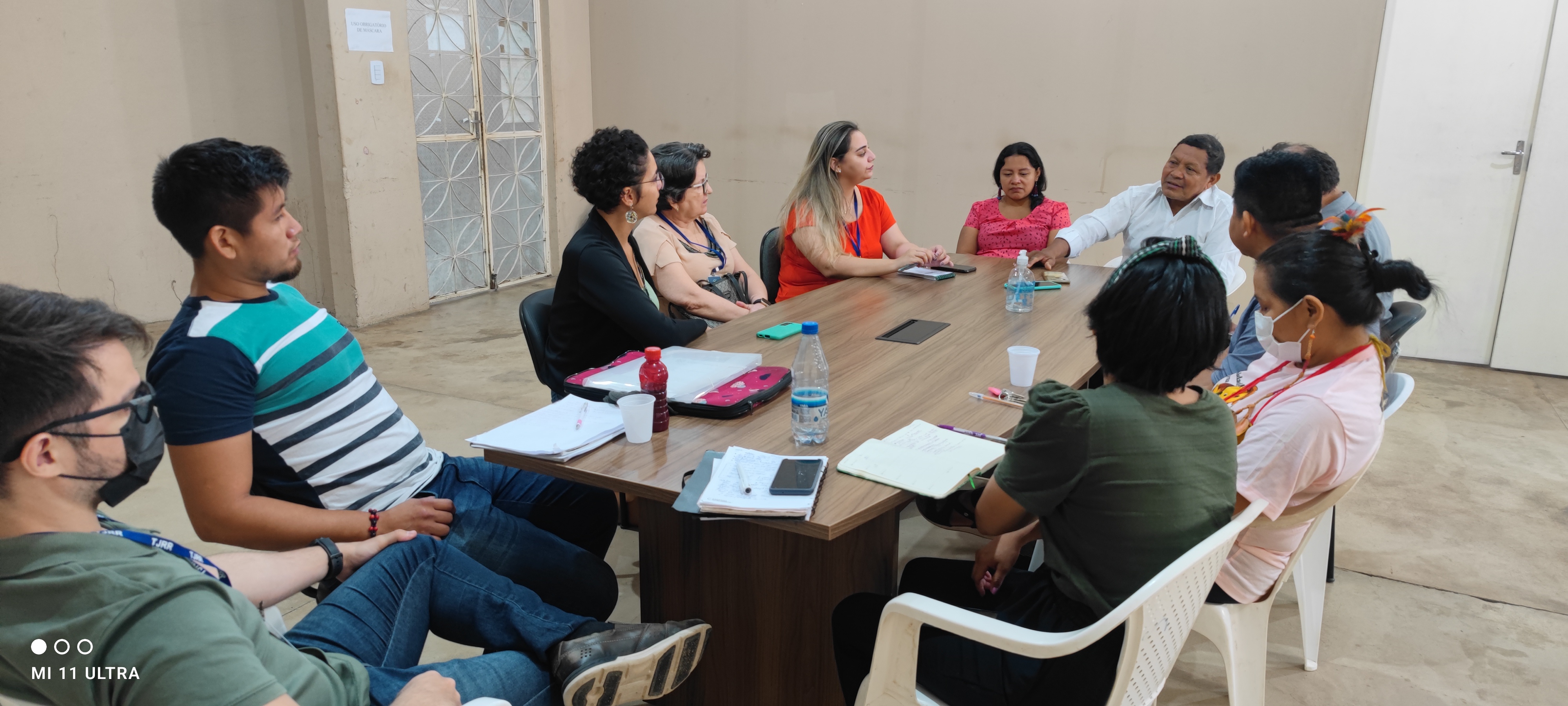 Imagem colorida retangular, mostra uma mesa com papéis para anotações e garrafas de água, ao redor estão 11 pessoas sentadas em cadeiras.