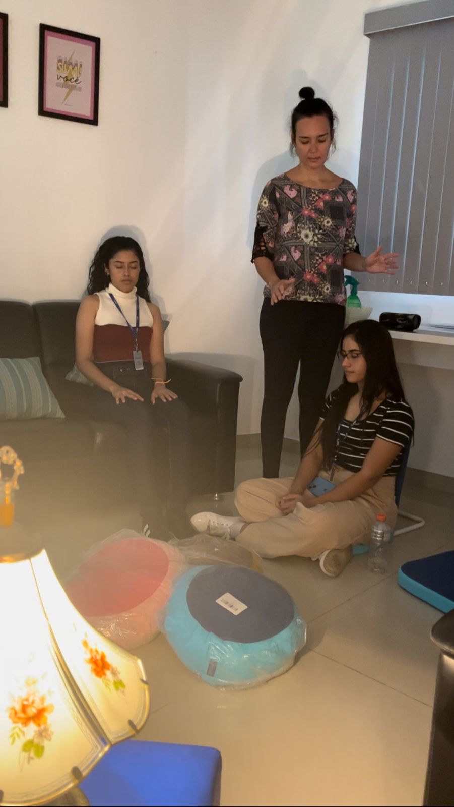 Imagem colorida mostrando 3 mulheres. Duas estão sentadas, de olhos fechados, meditando. A outra servidora está em pé, também de olhos fechados. No chão, a frente das servidoras tem almofadas coloridas. 