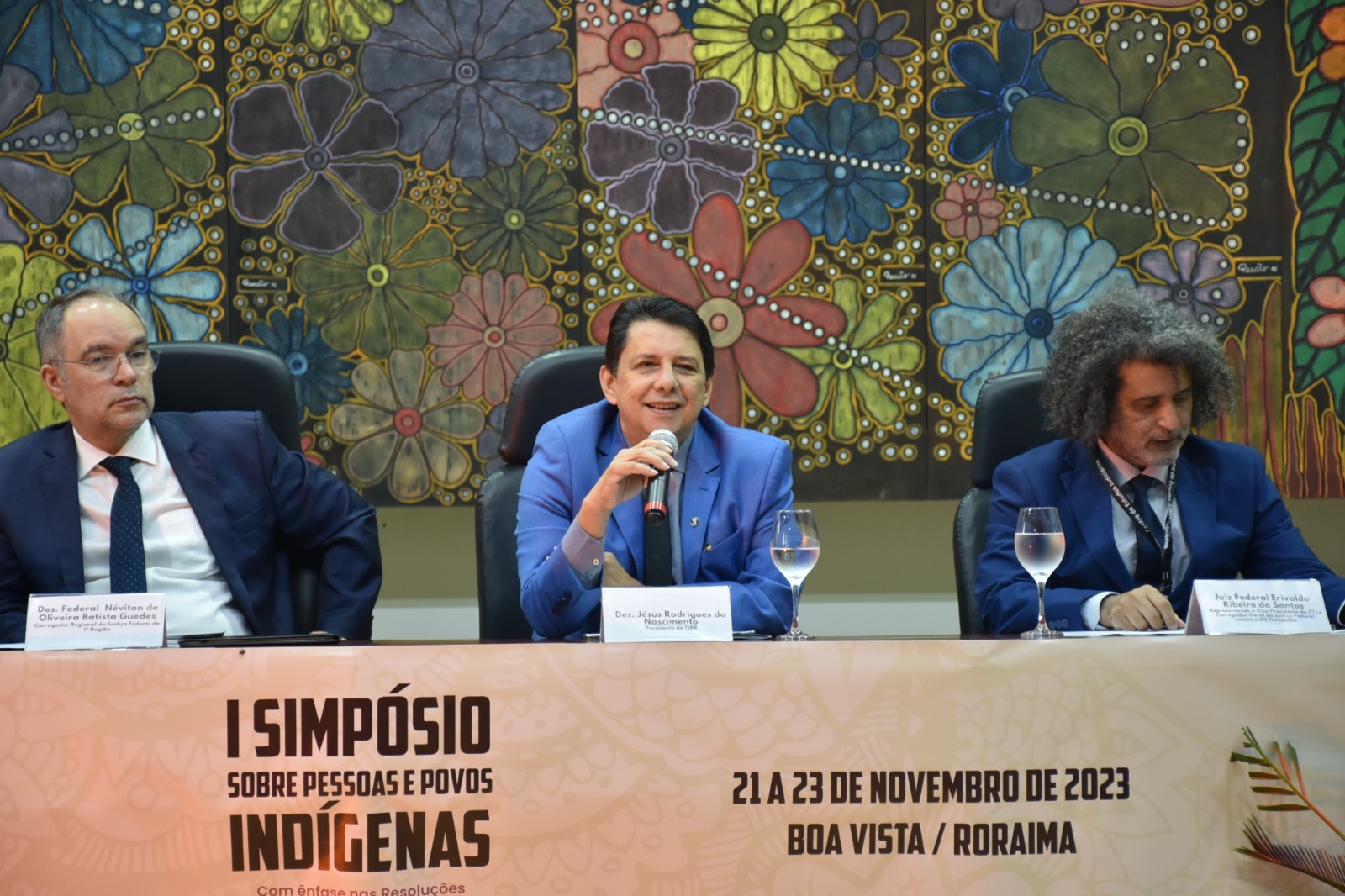  Foto colorida mostra o presidente do Poder Judiciário de Roraima, desembargador Jésus Nascimento, sentado com o microfone na mão, ao lado do  Corregedor Regional de Justiça Federal da 1ª Região, desembargador, Néviton Guedes e do o juiz federal Erivaldo Santos, o secretário nacional de acesso à justiça, Marivaldo Pereira.  No fundo há um quadro com diversas flores coloridas.