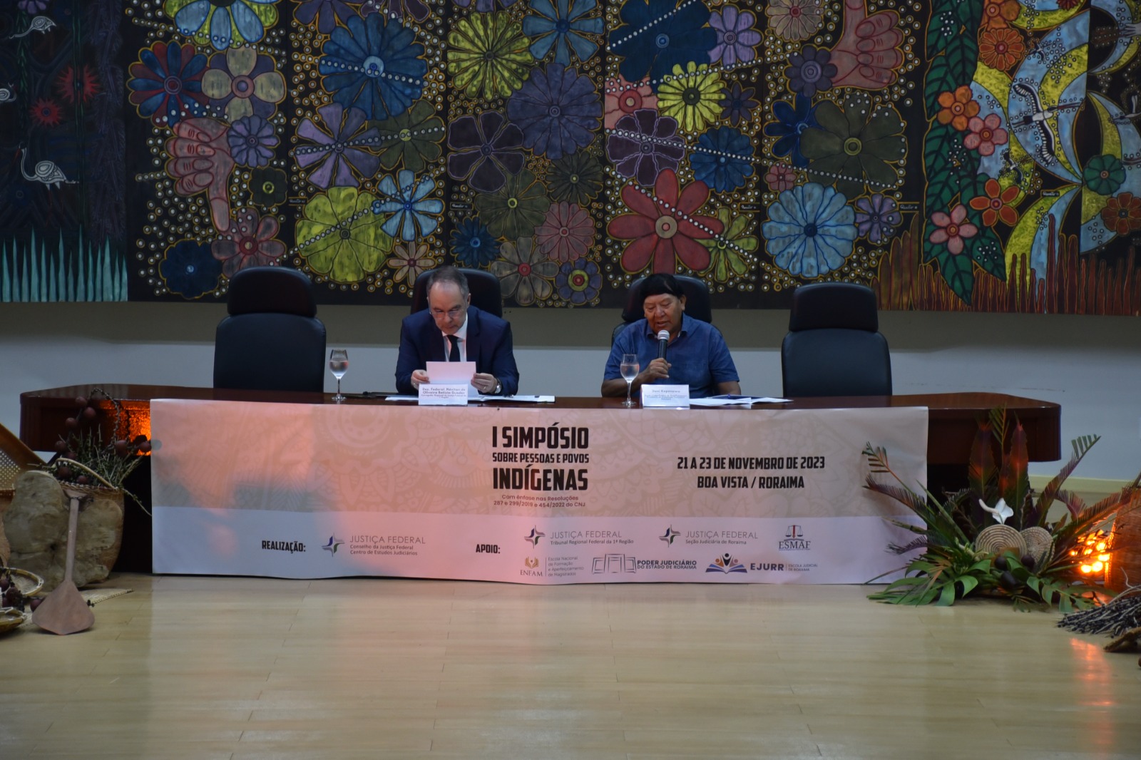  Foto colorida mostra o xamã e líder político do povo Yanomami, Davi Kopenawa, sentado com o microfone na mão, ao lado do  Corregedor Regional de Justiça Federal da 1ª Região, desembargador, Néviton Guedes, que está com um papel. No fundo há um quadro com diversas flores coloridas. A frente há uma faixa escrito “  I Simpósio Sobre Pessoas e Povos Indígenas”, “21 a 23 de novembro de 2023, Boa Vista / Roraima”.