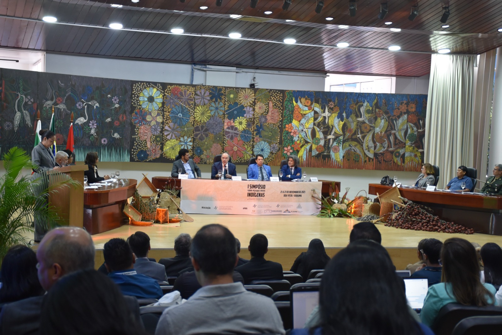 Foto colorida mostra uma plateia de costas para a câmera. A frente está o presidente do Poder Judiciário de Roraima, desembargador Jésus Nascimento, sentado ao lado do  Corregedor Regional de Justiça Federal da 1ª Região, desembargador Néviton Guedes, que está com o microfone na mão, o juiz federal Erivaldo Santos. No fundo há um quadro com diversas flores coloridas. A frente há uma faixa escrito “  I Simpósio Sobre Pessoas e Povos Indígenas”, “21 a 23 de novembro de 2023, Boa Vista / Roraima”.