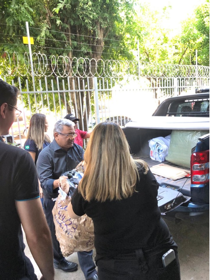 Foto colorida mostra um grupo com 5 pessoas, estando dois deles de costas para a câmera,  carregando a traseira de uma picape com materiais para doação.