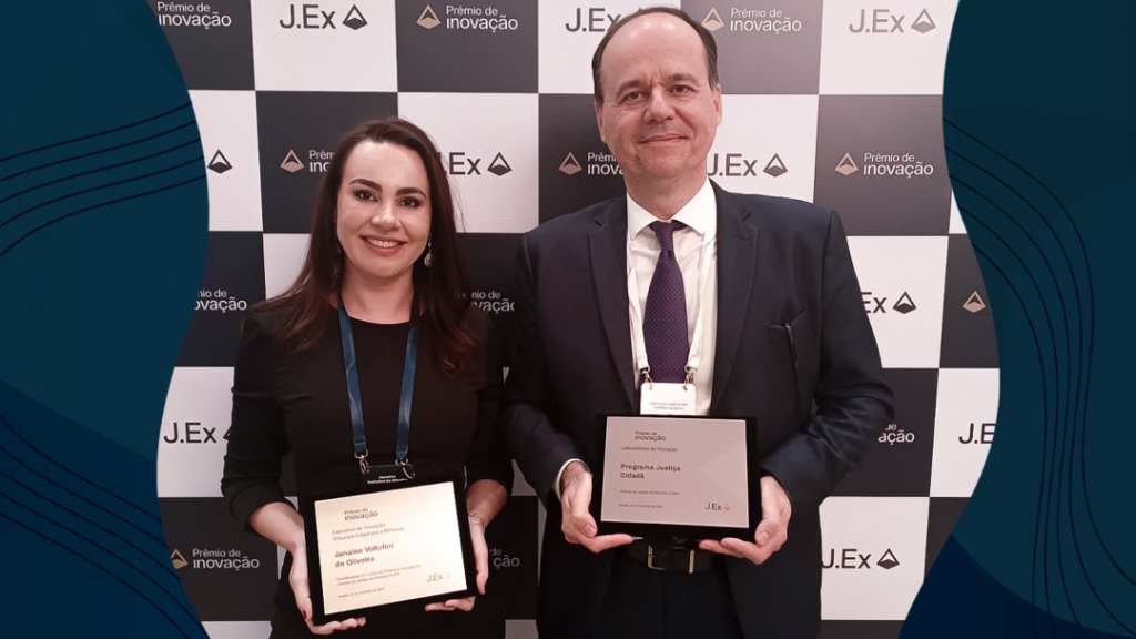 Foto colorida mostra o coordenador do Justiça Cidadã, desembargador Cristóvão Suter, posando para a foto ao lado da  coordenadora do Núcleo de Projetos e Inovação (NPI), Janaine Voltolini de Oliveira, ambos segurando placas de metal que simbolizam o prêmio  de inovação 2023.