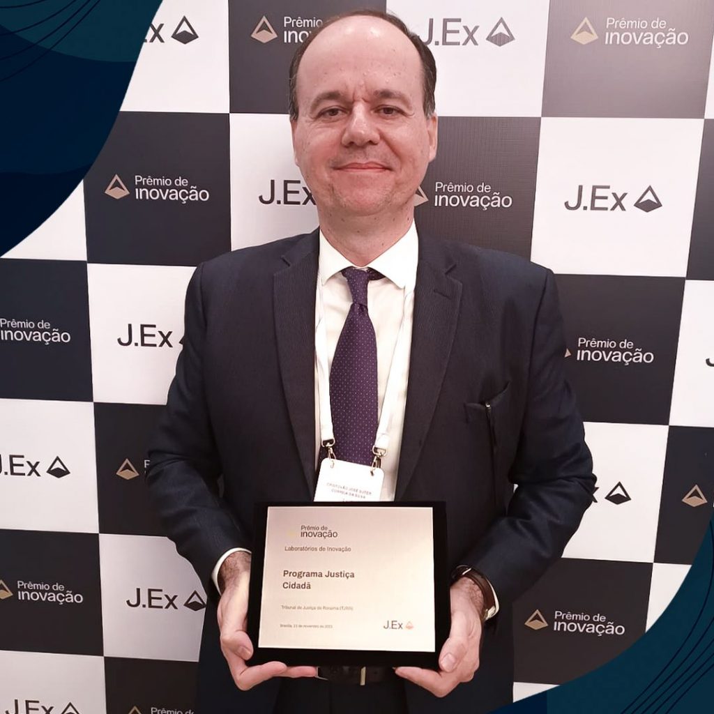 Imagem 3: Foto colorida mostra o coordenador do Justiça Cidadã, desembargador Cristóvão Suter, posando para a foto segurando placas de metal que simbolizam o prêmio  de inovação 2023.