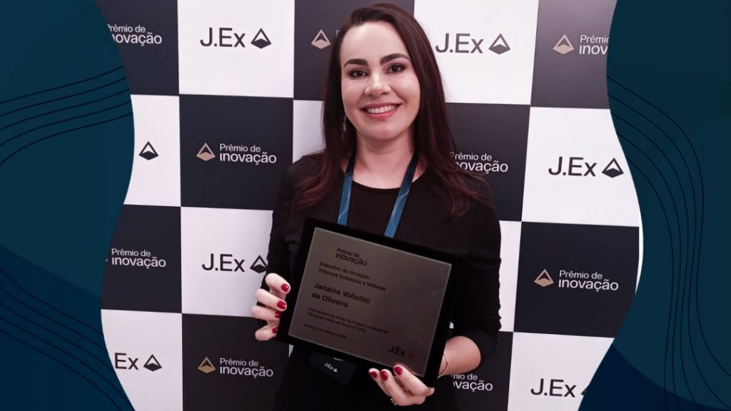 oto colorida mostra a  coordenadora do Núcleo de Projetos e Inovação (NPI), Janaine Voltolini de Oliveira, posando para a foto segurando placas de metal que simbolizam o prêmio  de inovação 2023.