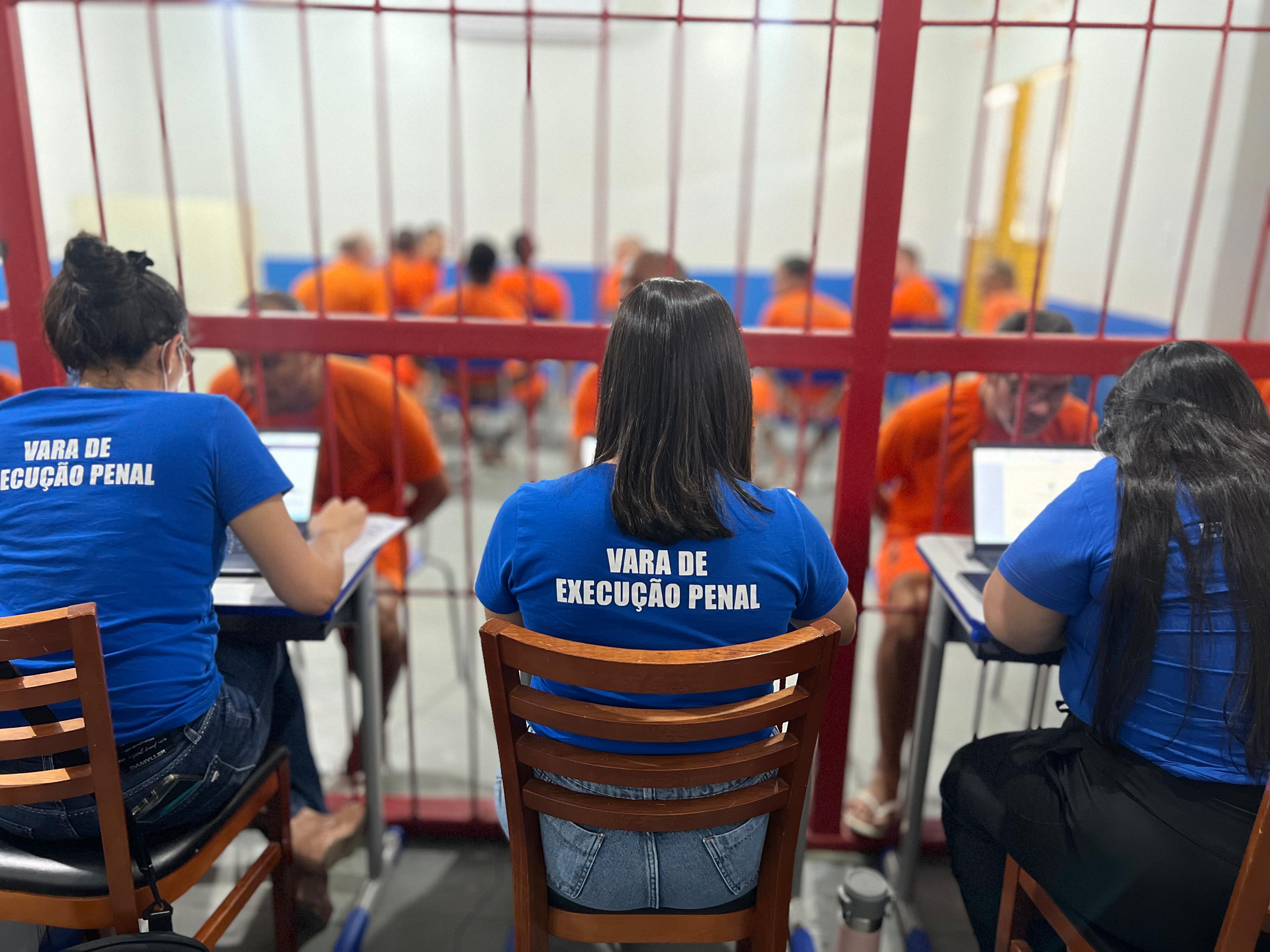 foto colorida de costa na cadeia masculina com tres servioras do tjrr sentadas realizando atendimento em frente das celas 